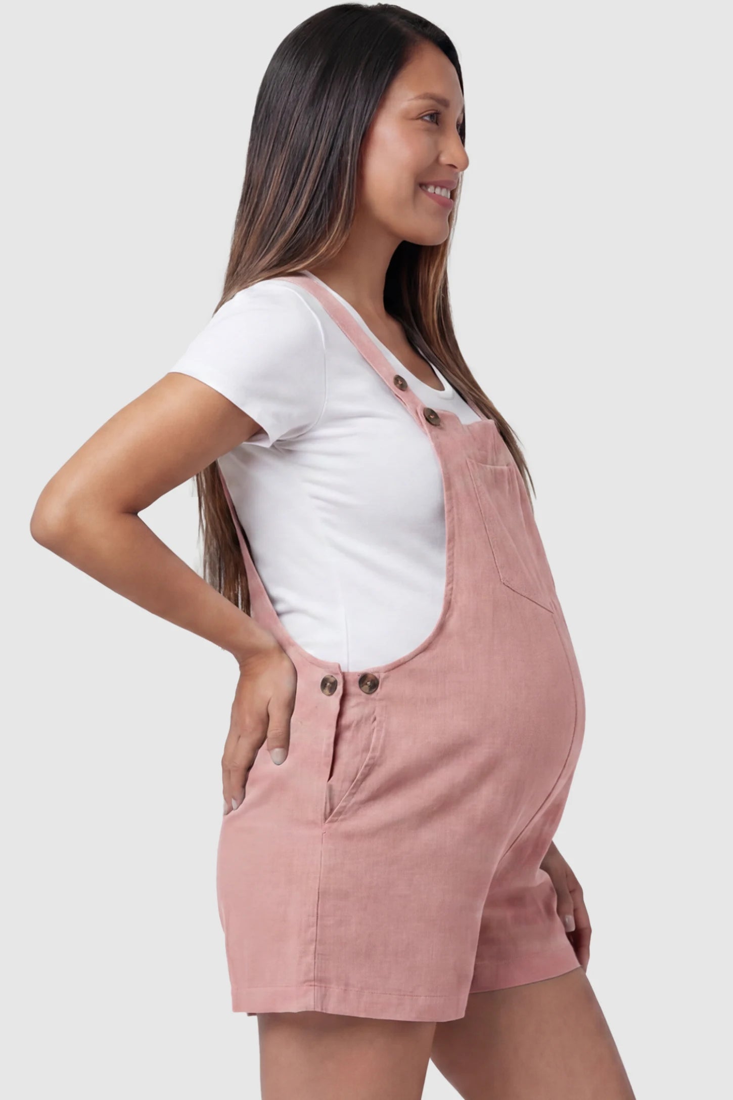 Adjustable Waist Pocketed Maternity Short Overalls Adjustable Waist Pocketed Maternity Short Overalls in Pink featuring functional side buttons on a pregnant model, side view.