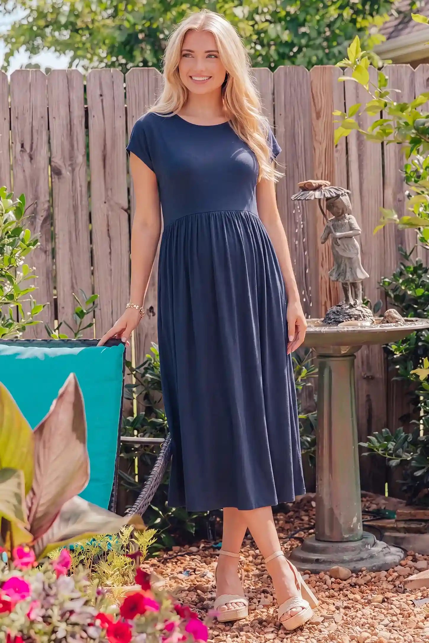 Crew Neck Short Sleeve Maternity Midi Dress Crew Neck Short Sleeve Maternity Midi Dress in Navy blue featuring a flowy shift silhouette on a pregnant woman with sunglasses standing on a city sidewalk next to green plants, side view.