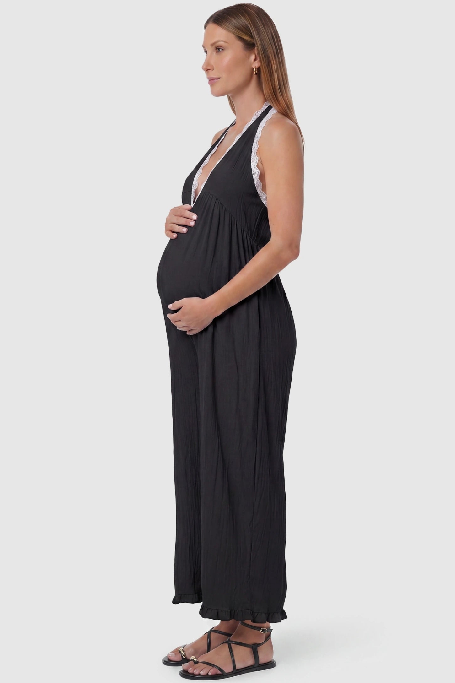 Deep V-Neck Lace Trim Wide Leg Maternity Jumpsuit Deep V-Neck Lace Trim Wide Leg Maternity Jumpsuit in Black featuring a flowy wide leg silhouette with lace trim detail on a pregnant model, side view.