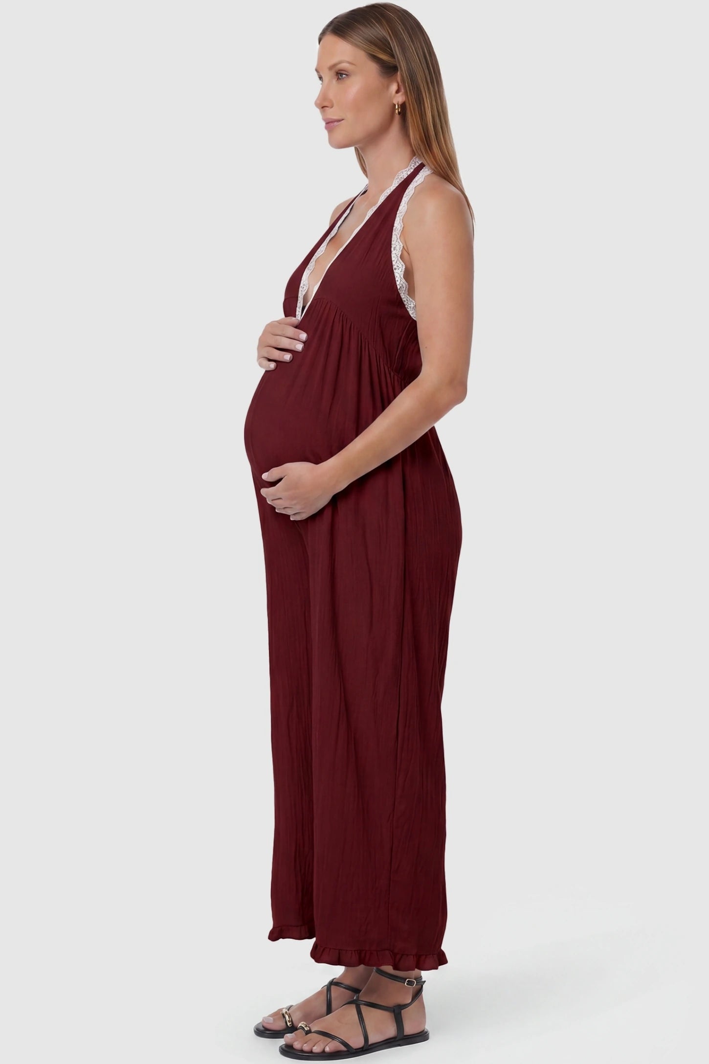 Deep V-Neck Lace Trim Wide Leg Maternity Jumpsuit Deep V-Neck Lace Trim Wide Leg Maternity Jumpsuit in Burgundy featuring a flowy wide leg silhouette with lace trim detail on a pregnant model, side view.