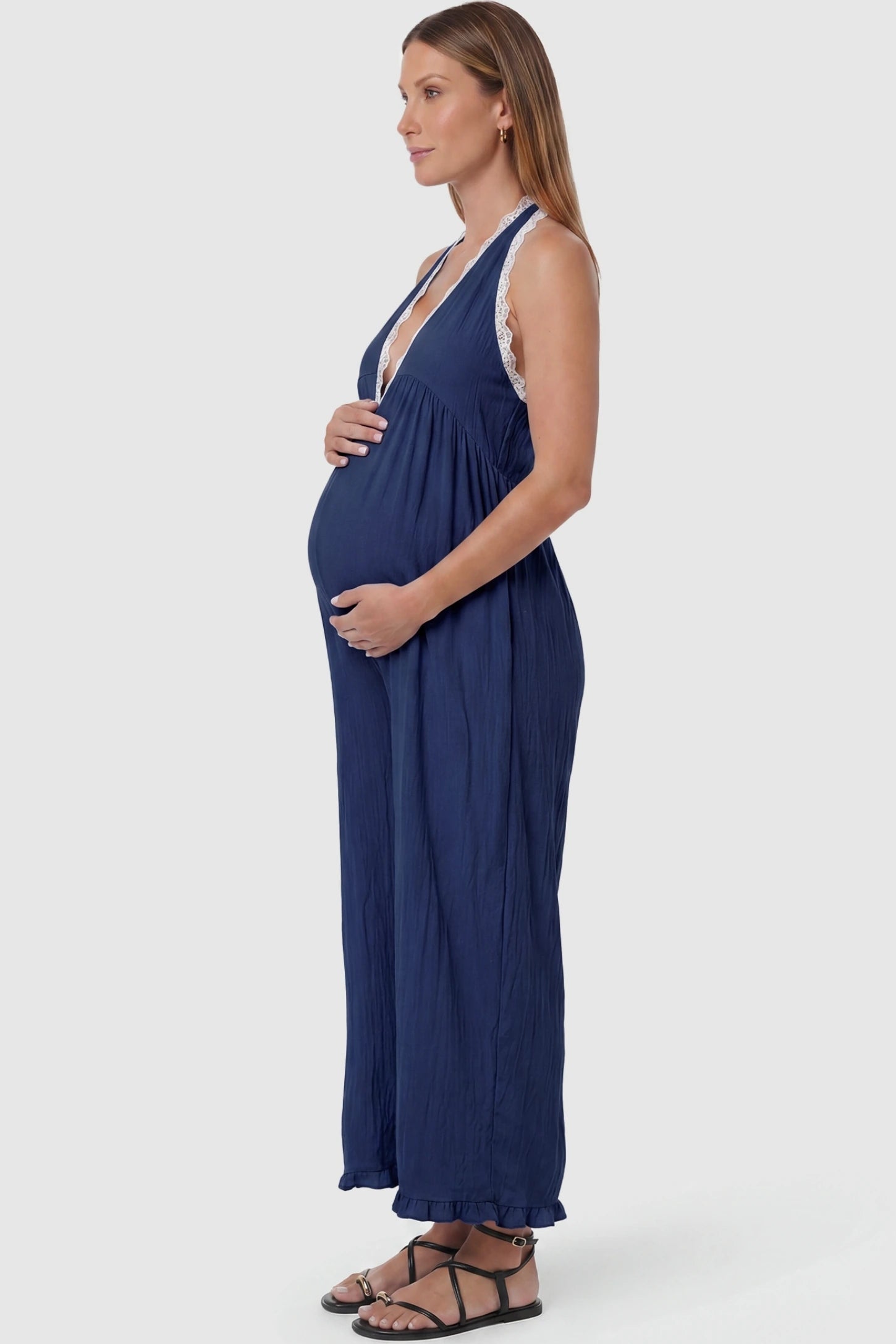 Deep V-Neck Lace Trim Wide Leg Maternity Jumpsuit Deep V-Neck Lace Trim Wide Leg Maternity Jumpsuit in Navy Blue featuring a flowy wide leg silhouette with lace trim detail on a pregnant model, side view.