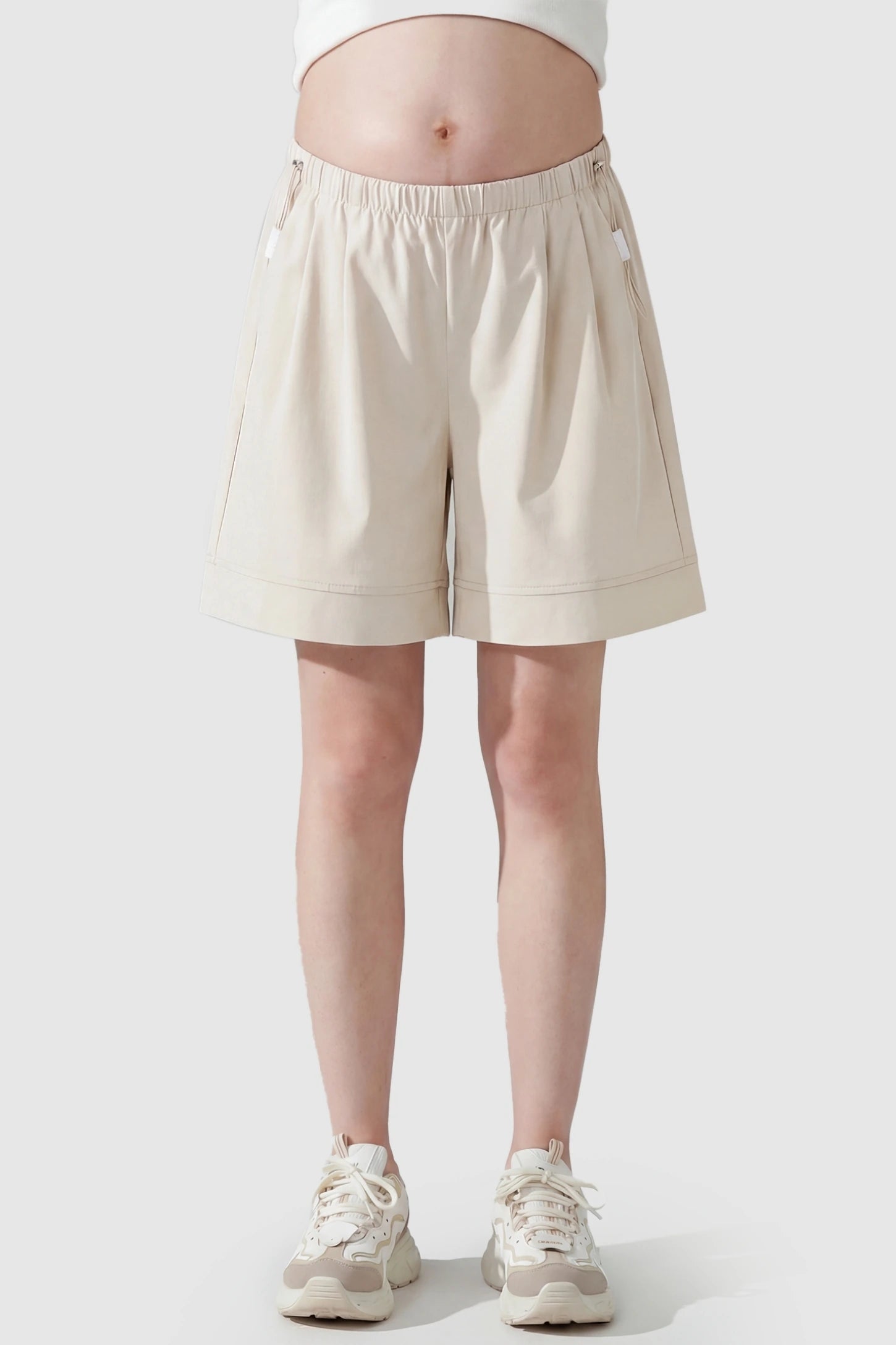 Pocketed Side Drawstring Maternity Shorts Beige Elasticated Waist Wide Leg Maternity Shorts featuring a flexible tummy band on a pregnant model, front view.