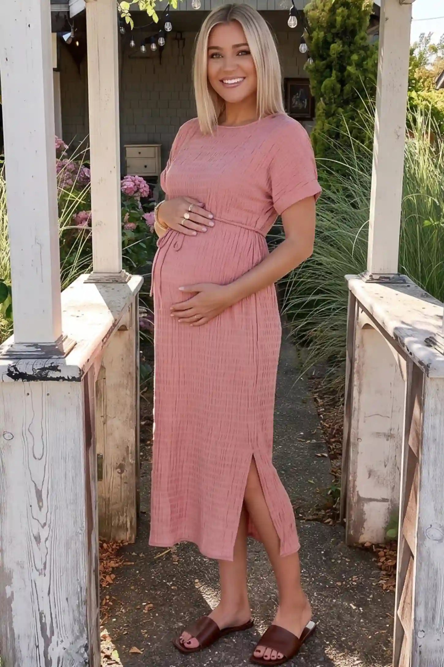 Empire Waist Round Neck Maternity Midi Dress in Pink featuring elasticated waistband with a decorative tie detail on a plus size pregnant woman in outdoor setting, side view.