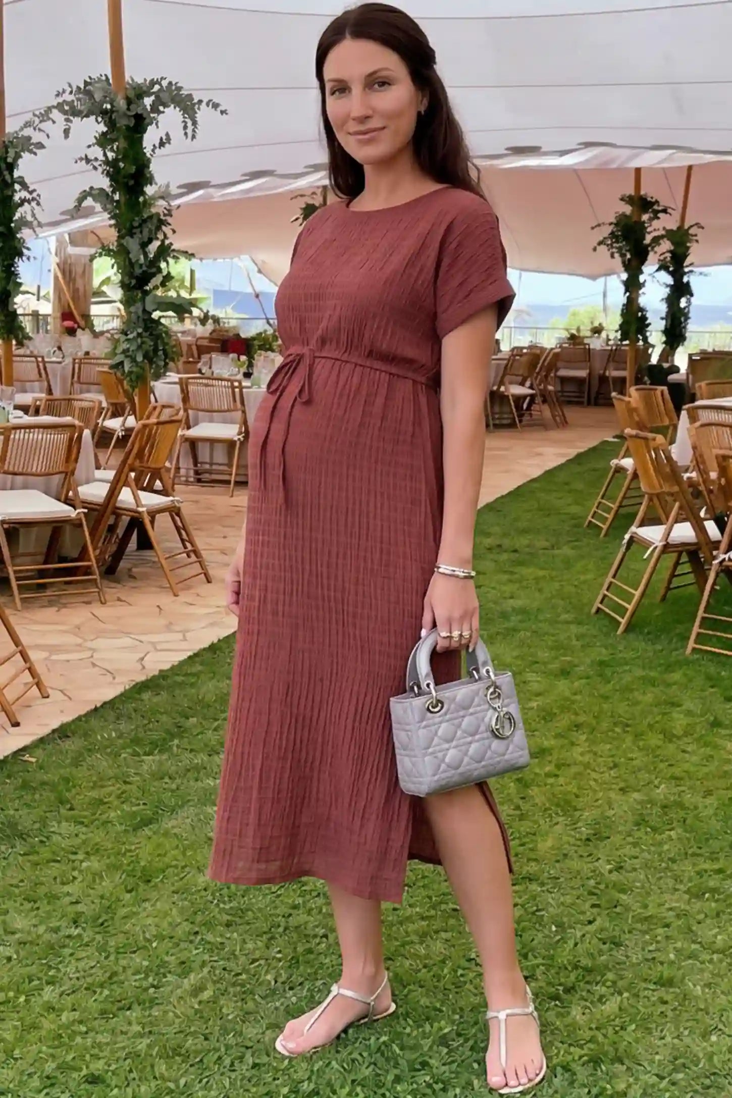 Empire Waist Round Neck Maternity Midi Dress Empire Waist Round Neck Maternity Midi Dress in Wine Red featuring elasticated waistband with a decorative tie detail on a plus size pregnant woman in outdoor setting, front view.