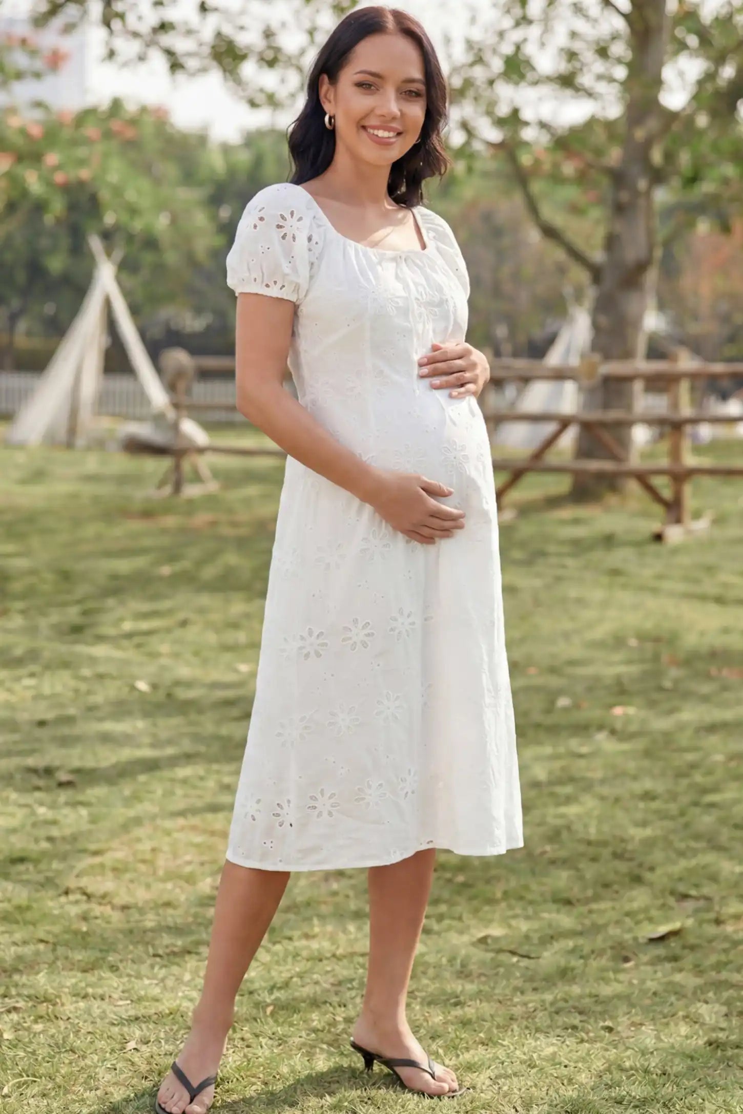 Eyelet Embroidered Short Sleeve Maternity Midi Dress Eyelet Embroidered Short Sleeve Maternity Midi Dress in white featuring a square neckline and drawstring waist on a pregnant model in a park, front view.