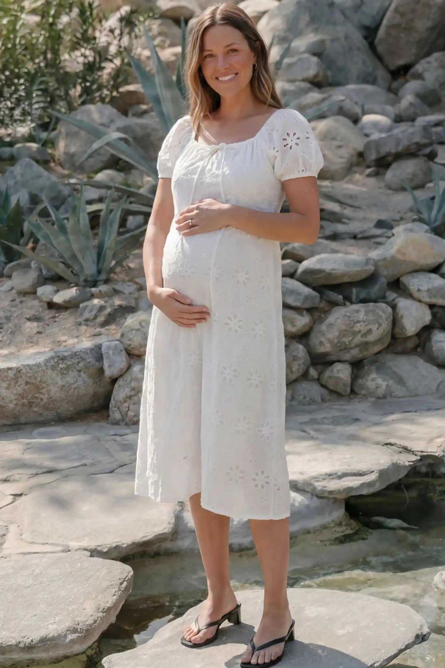 Eyelet Embroidered Short Sleeve Maternity Midi Dress Eyelet Embroidered Short Sleeve Maternity Midi Dress in white featuring a square neckline and drawstring waist on a pregnant model by a stone garden, front view.