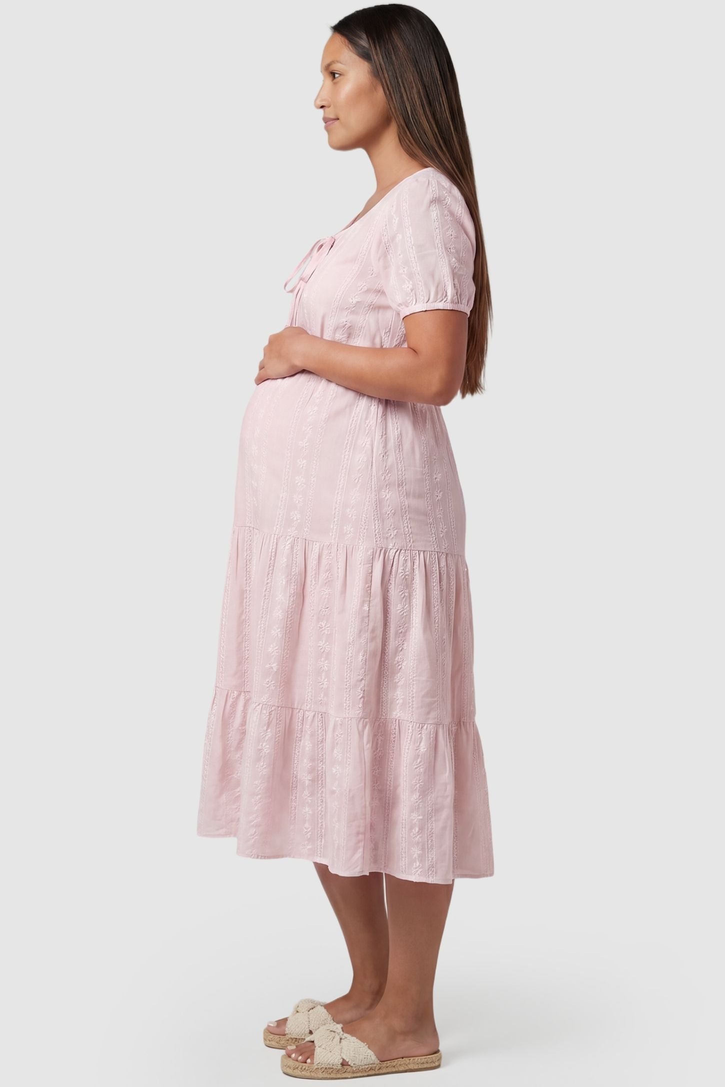 Eyelet Scoop Neck Tiered Maternity Dress Eyelet Scoop Neck Tiered Maternity Dress in Pink featuring a tiered flounce skirt on a pregnant model, side view.