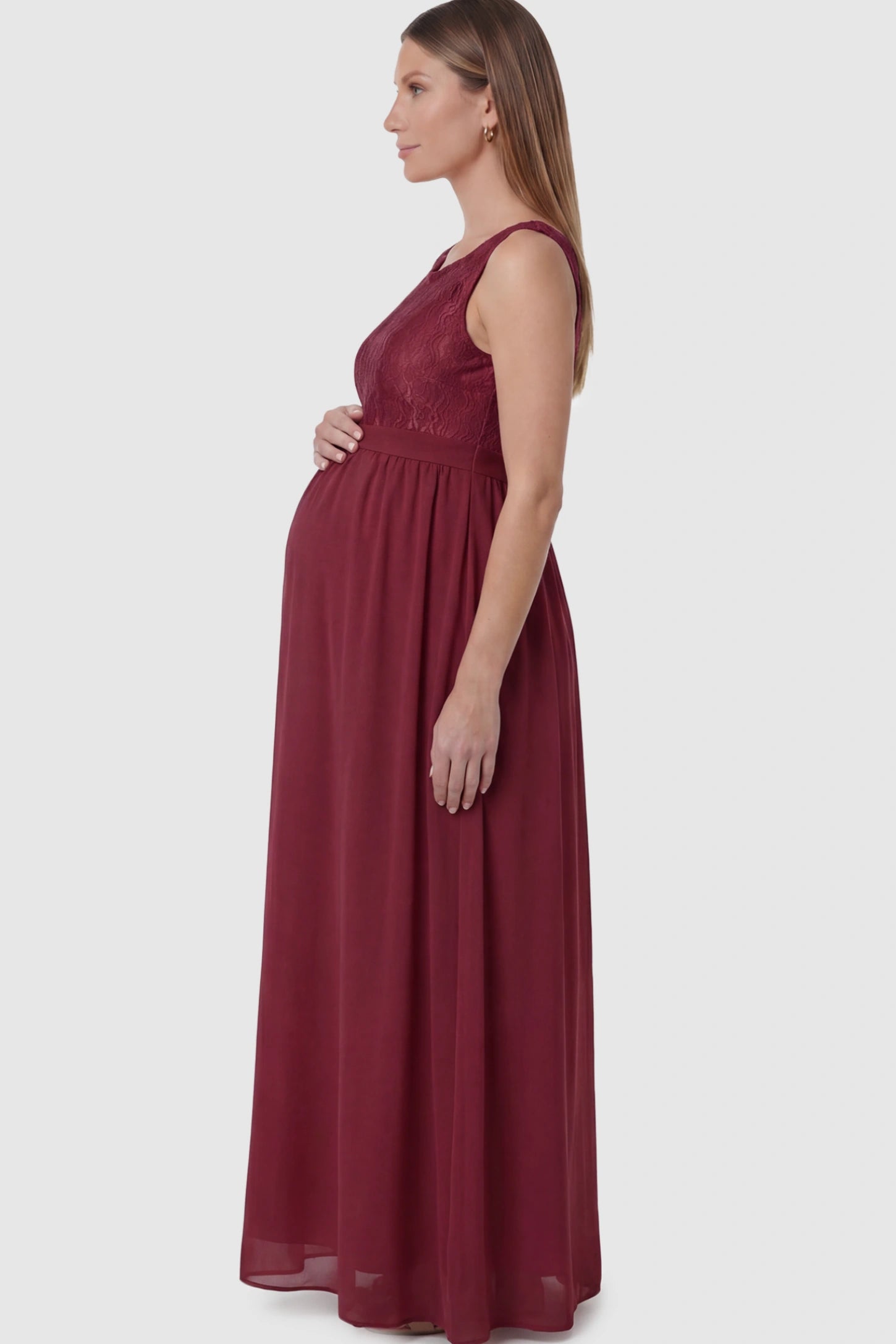 Floral Lace Sleeveless Maternity Maxi Dress Floral Lace Sleeveless Maternity Maxi Dress in Burgundy featuring a flowing skirt on a pregnant model showing side.