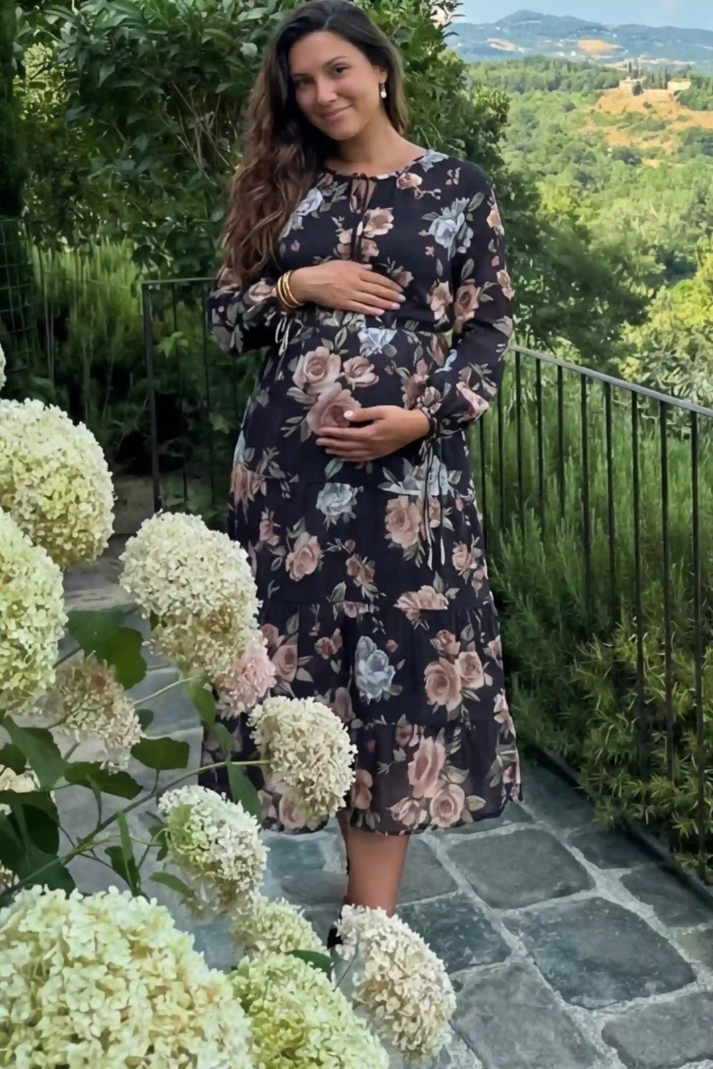 Floral Print Tiered Maternity Midi Dress Floral Print Tiered Midi Maternity Dress in dark floral featuring adjustable tie-cuff sleeves on a pregnant woman.