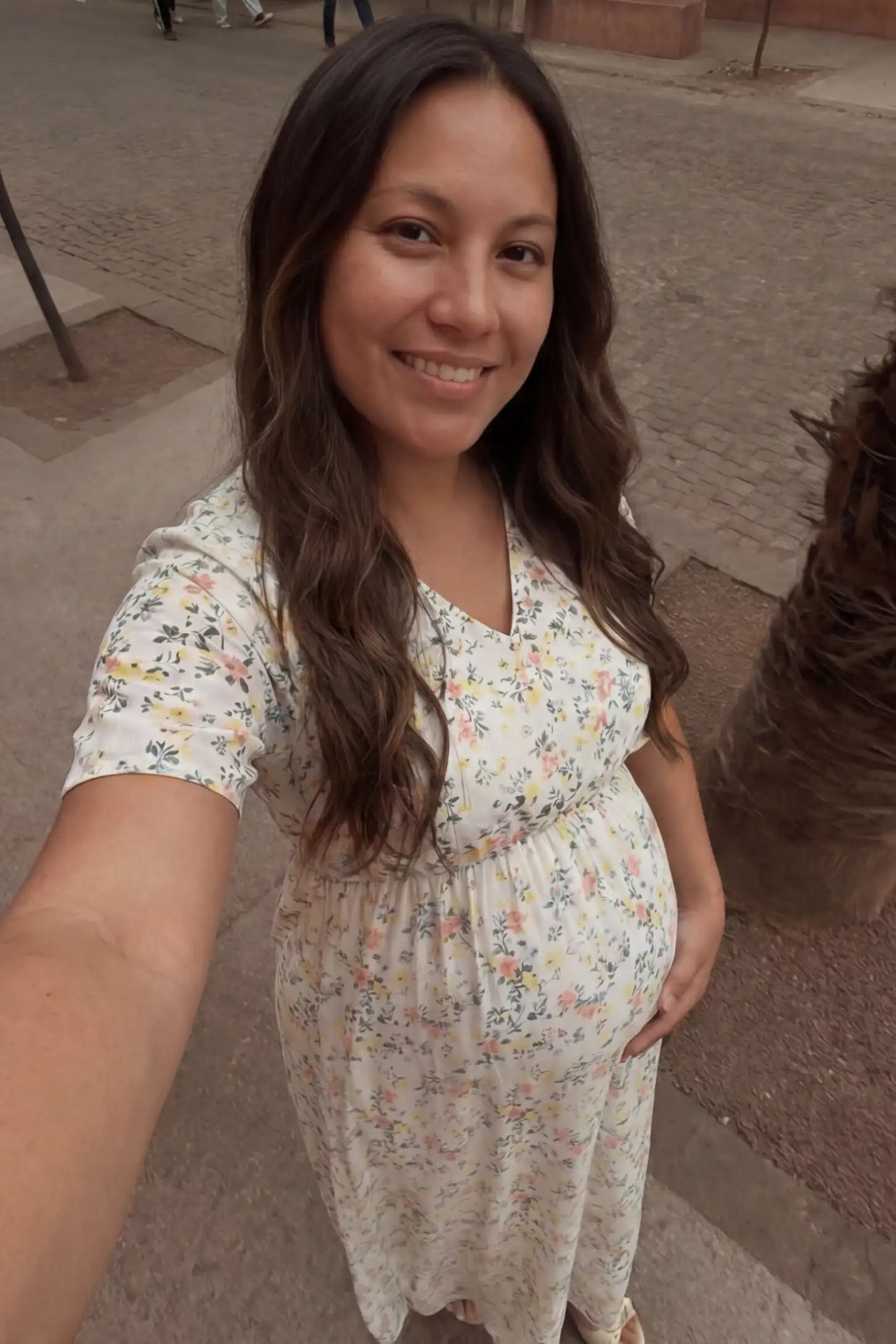 Floral V-Neck Side-Slit Maternity Maxi Dress Floral V-Neck Side-Slit Maternity Maxi Dress in white with a pastel ditsy floral pattern and an empire waist, shown from a high-angle selfie perspective by a pregnant woman, front view.