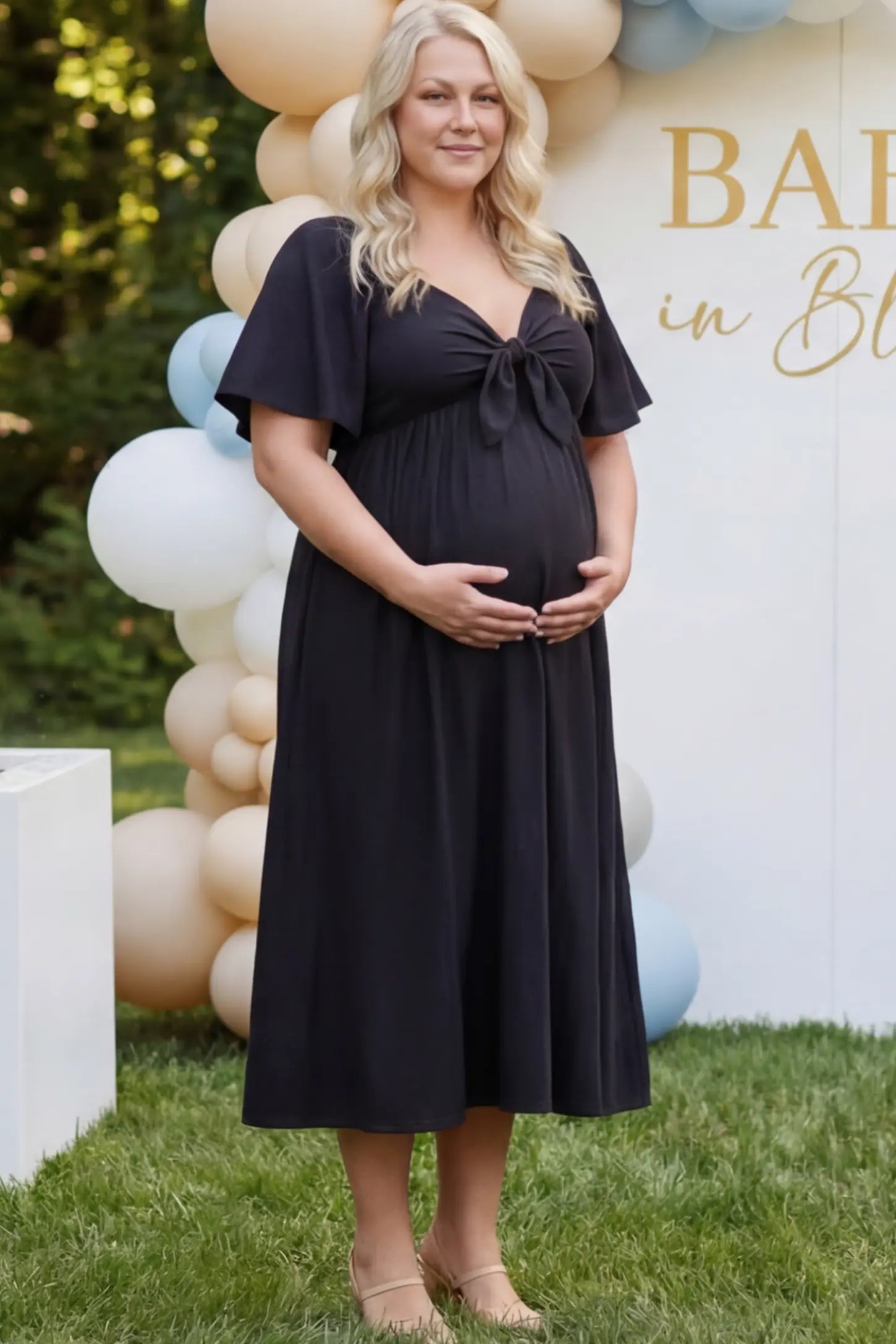 Front Tie Flutter Sleeve Maternity Midi Dress Front Tie Flutter Sleeve Maternity Midi Dress in Black featuring a knotted V-neckline and flowy skirt on a pregnant woman.