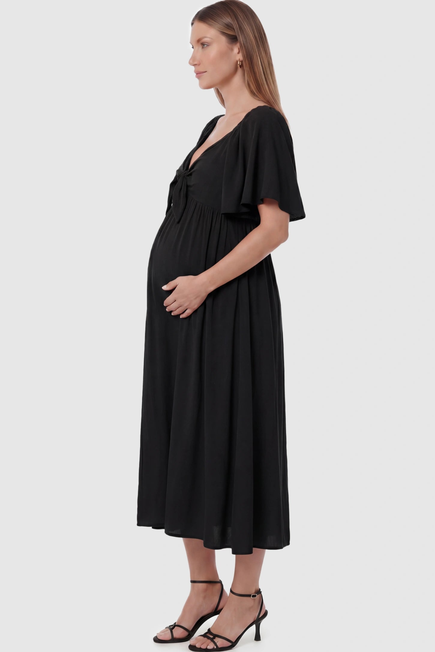 Front Tie Flutter Sleeve Maternity Midi Dress Front Tie Flutter Sleeve Maternity Midi Dress in Black featuring a knotted V-neckline and flowy skirt on a pregnant model showing side.