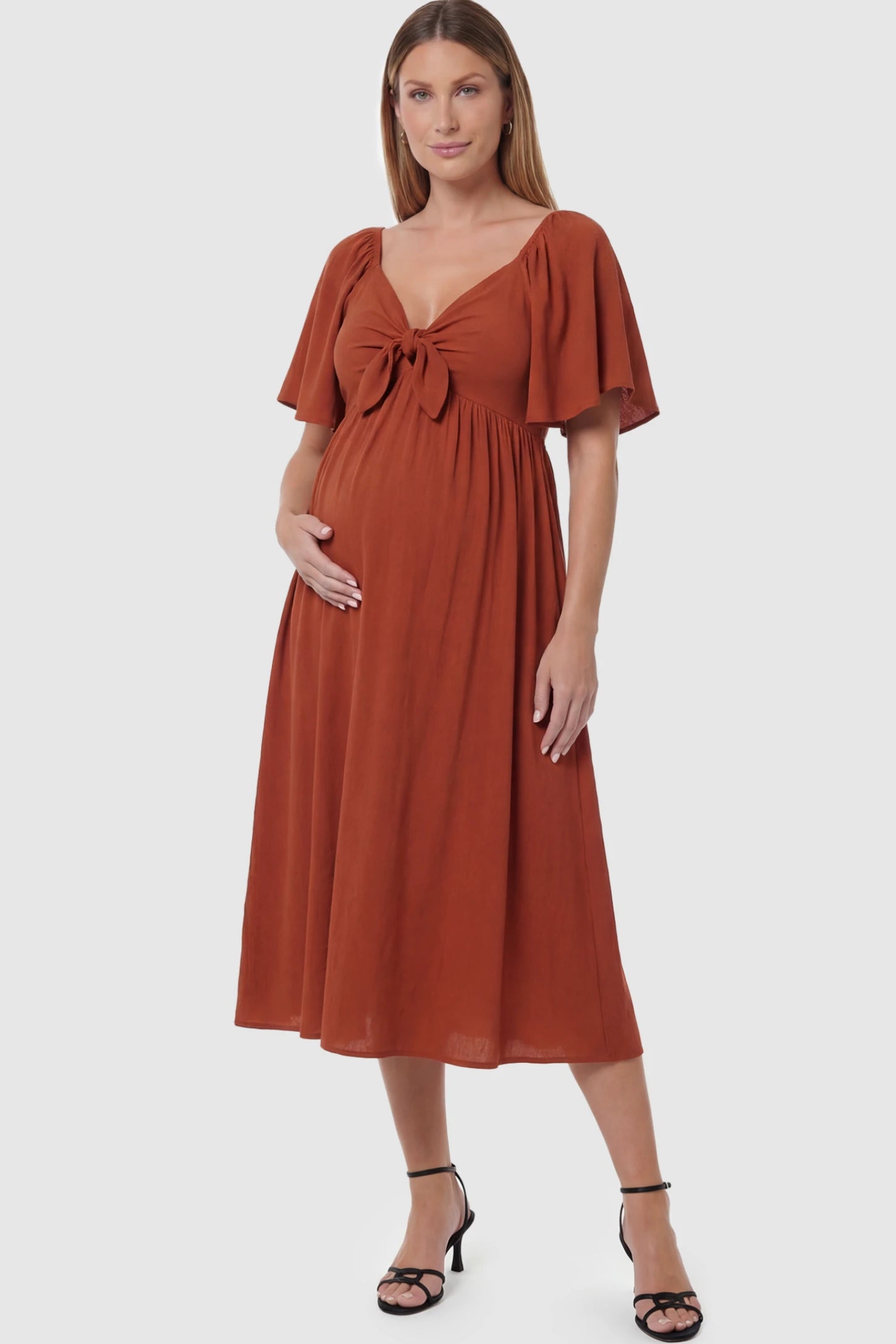 Front Tie Flutter Sleeve Maternity Midi Dress Front Tie Flutter Sleeve Maternity Midi Dress in Brick Red featuring a knotted V-neckline and flowy skirt on a pregnant model posing front.