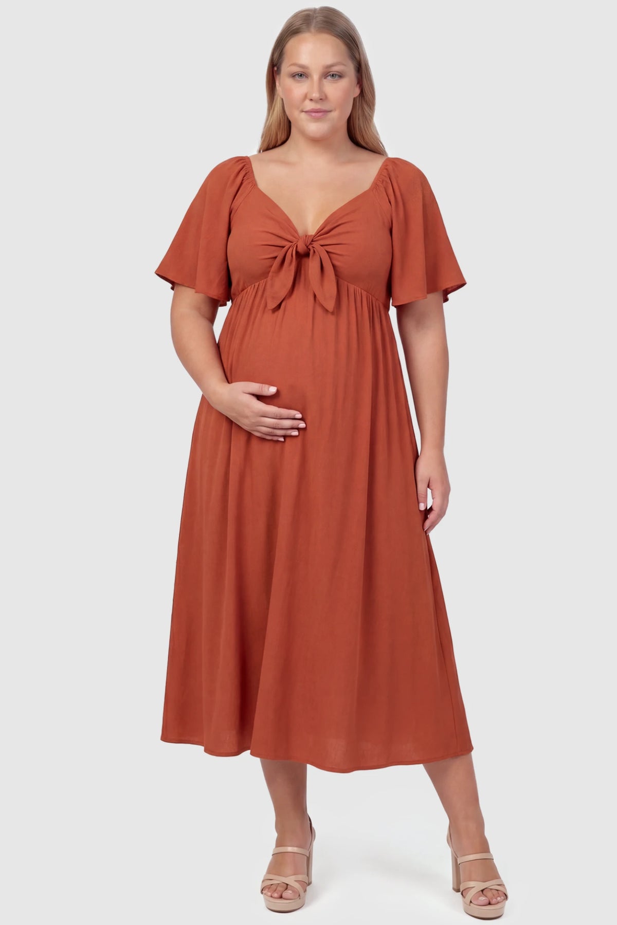 Front Tie Flutter Sleeve Maternity Midi Dress Front Tie Flutter Sleeve Maternity Midi Dress in brick red featuring a knotted V-neckline and flowy skirt on a plus size pregnant model, front view.