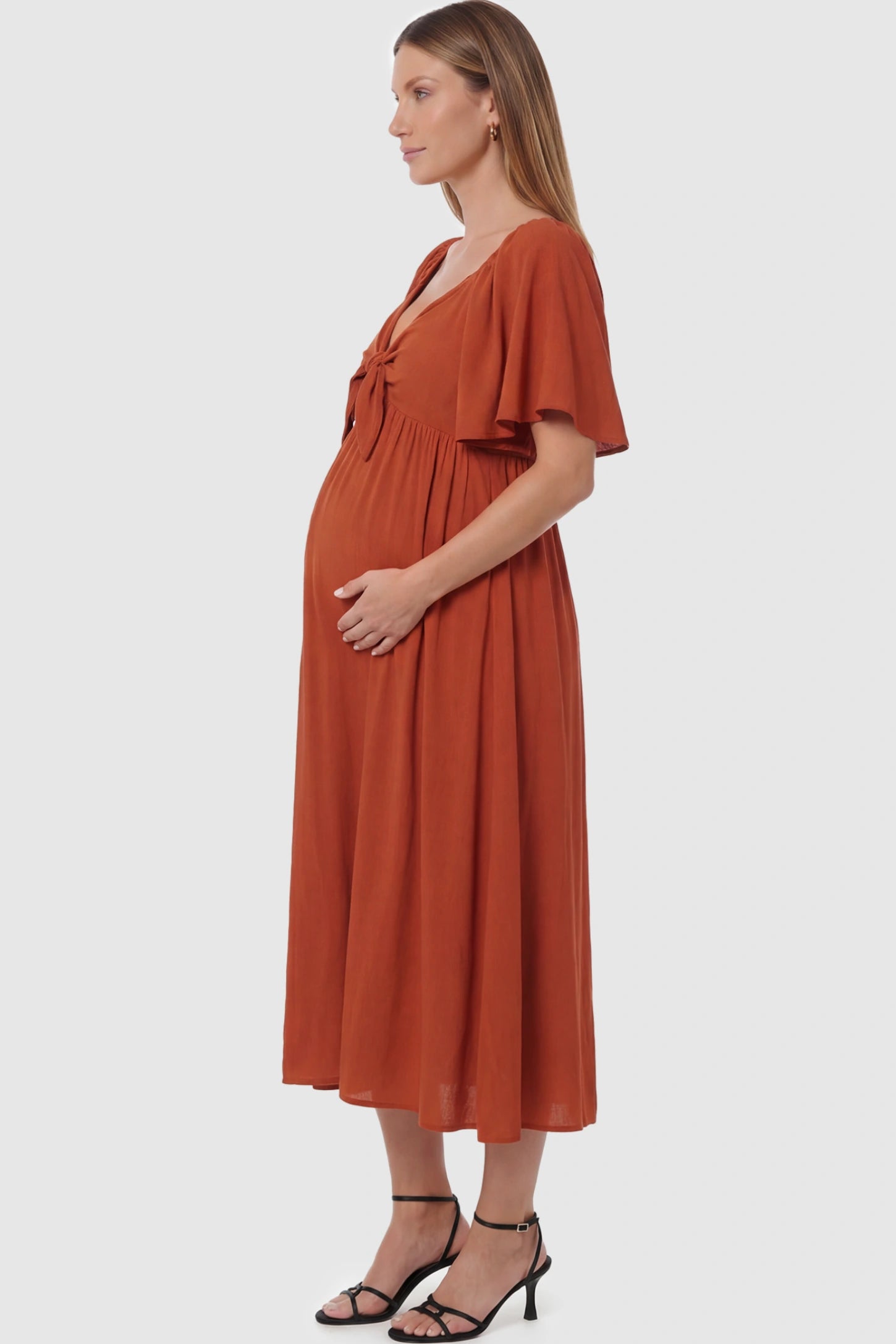 Front Tie Flutter Sleeve Maternity Midi Dress Front Tie Flutter Sleeve Maternity Midi Dress in Brick Red featuring a knotted V-neckline and flowy skirt on a pregnant model showing side.