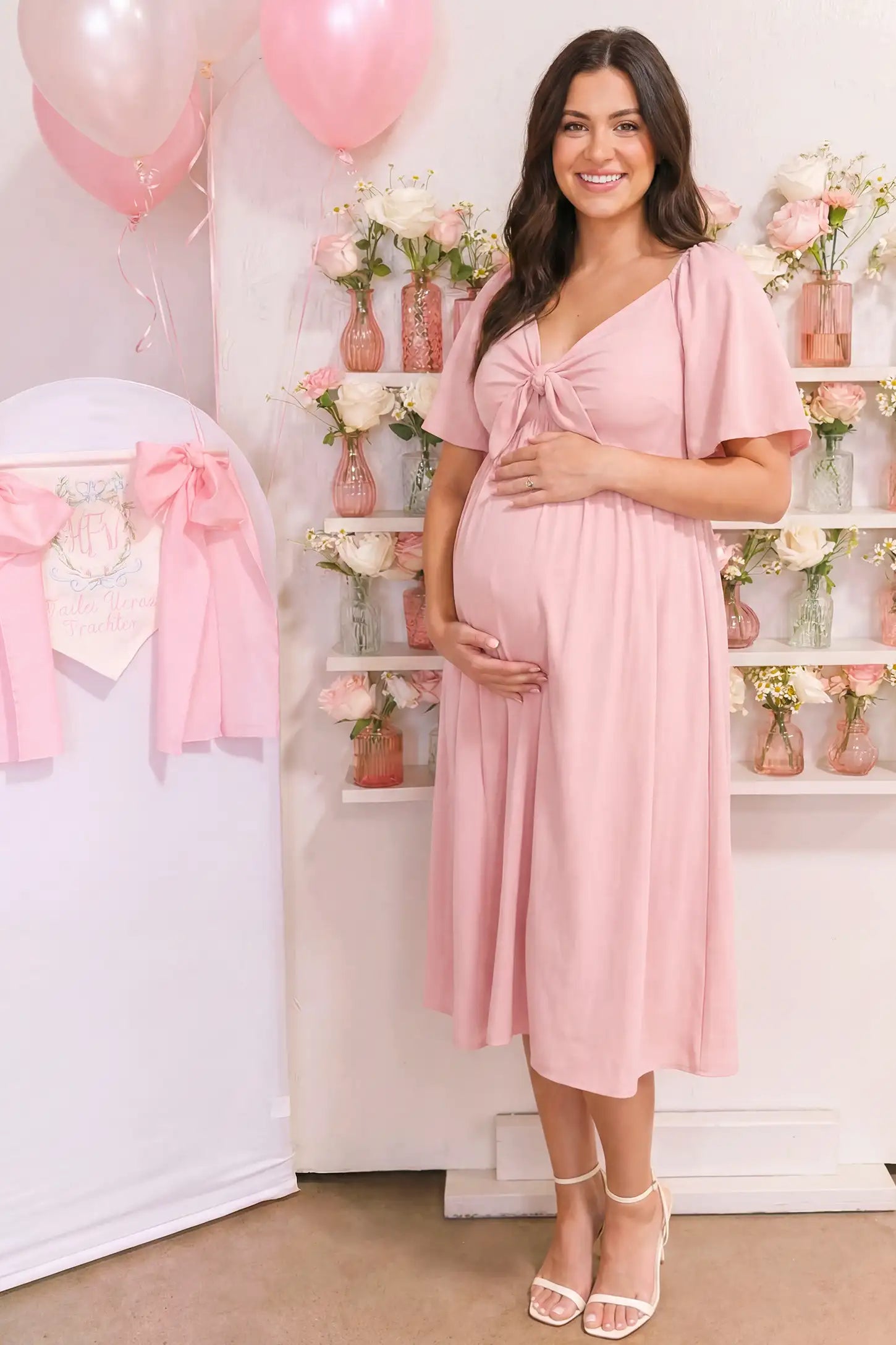 Front Tie Flutter Sleeve Maternity Midi Dress in pink featuring a knotted V-neckline and flowy skirt on a pregnant woman posing front.