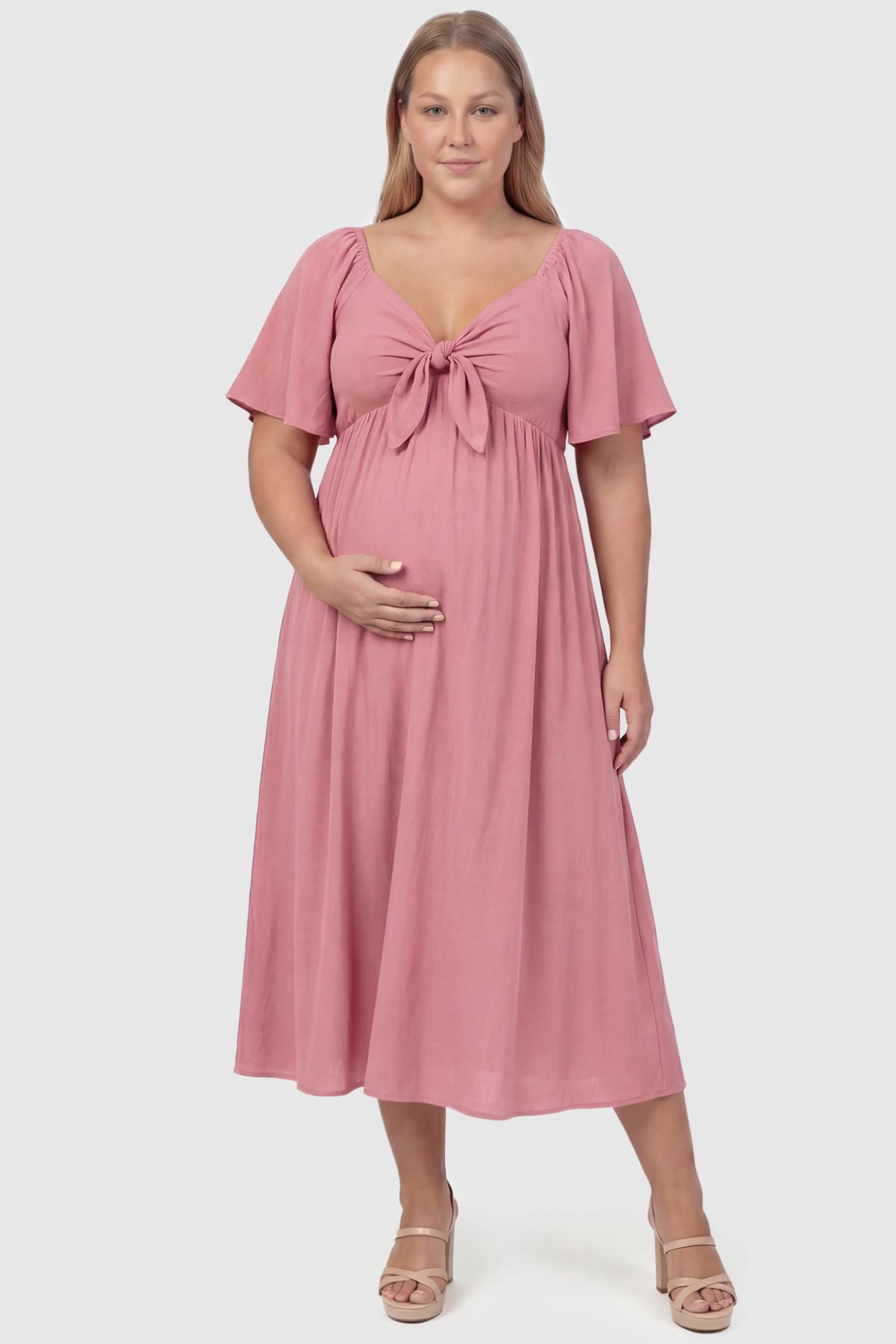 Front Tie Flutter Sleeve Maternity Midi Dress Front Tie Flutter Sleeve Maternity Midi Dress in pink featuring a knotted V-neckline and flowy skirt on a plus size pregnant model, front view.
