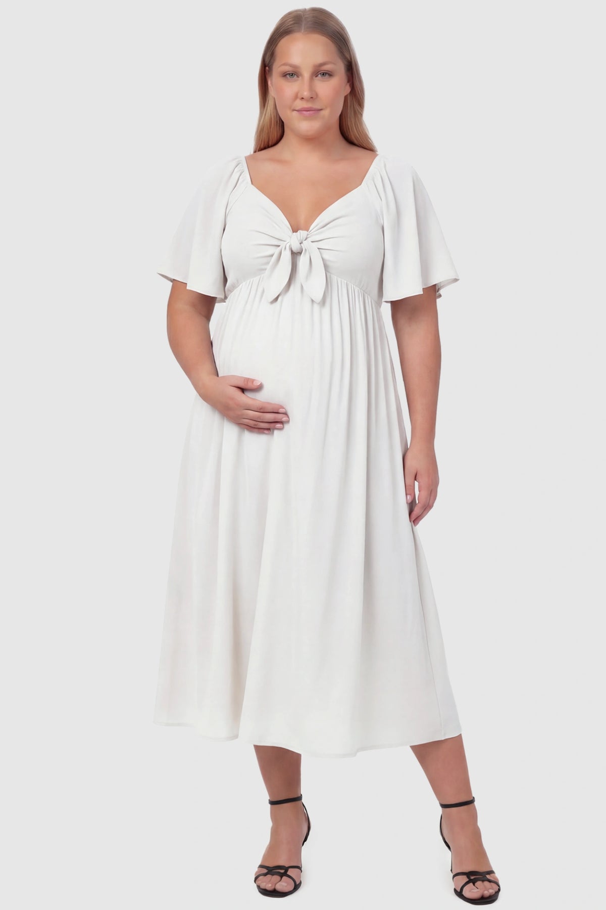Front Tie Flutter Sleeve Maternity Midi Dress Front Tie Flutter Sleeve Maternity Midi Dress in white featuring a knotted V-neckline and flowy skirt on a plus size pregnant model, front view.