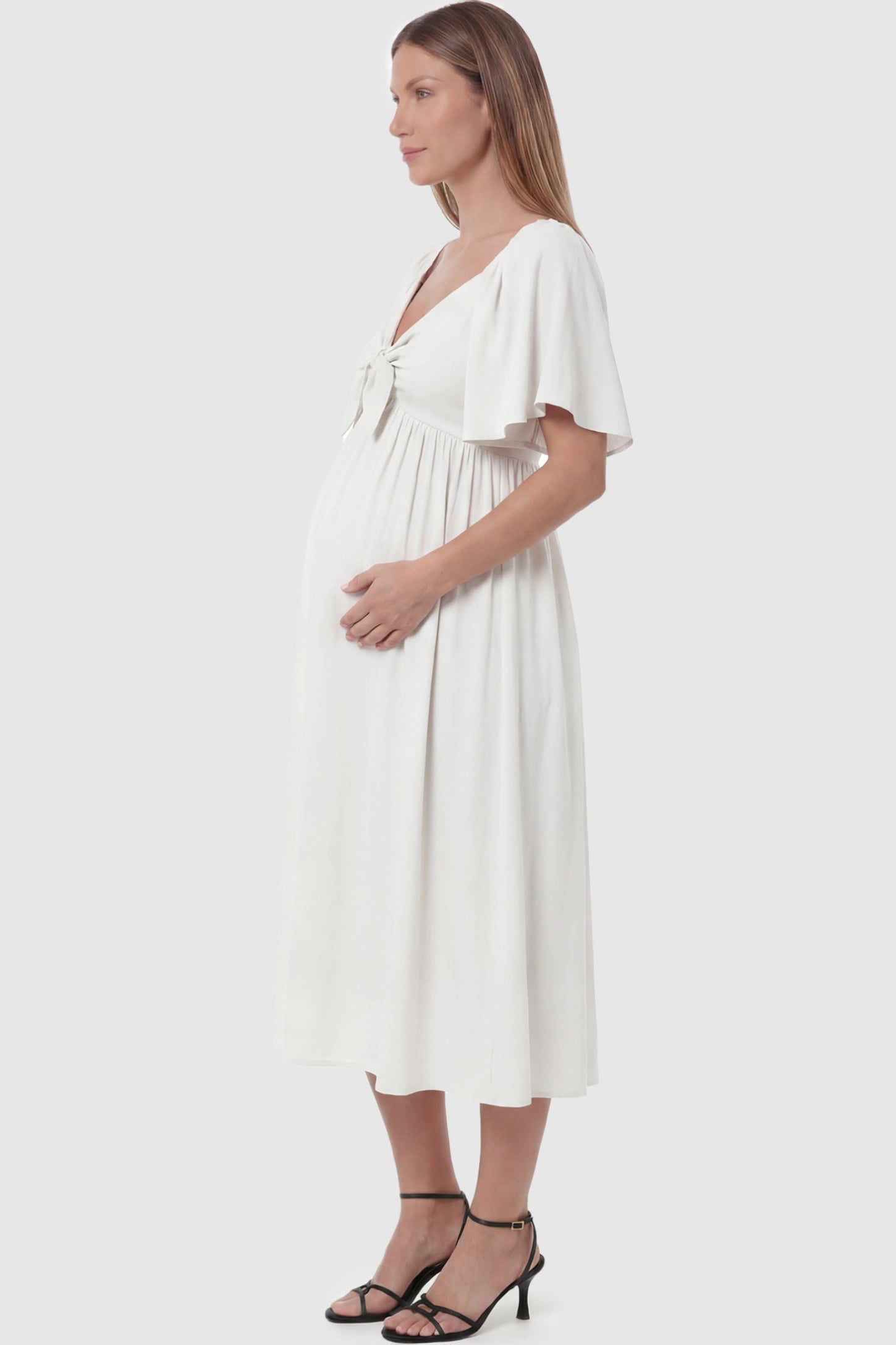Front Tie Flutter Sleeve Maternity Midi Dress Front Tie Flutter Sleeve Maternity Midi Dress in white featuring a knotted V-neckline and flowy skirt on a pregnant model showing side.