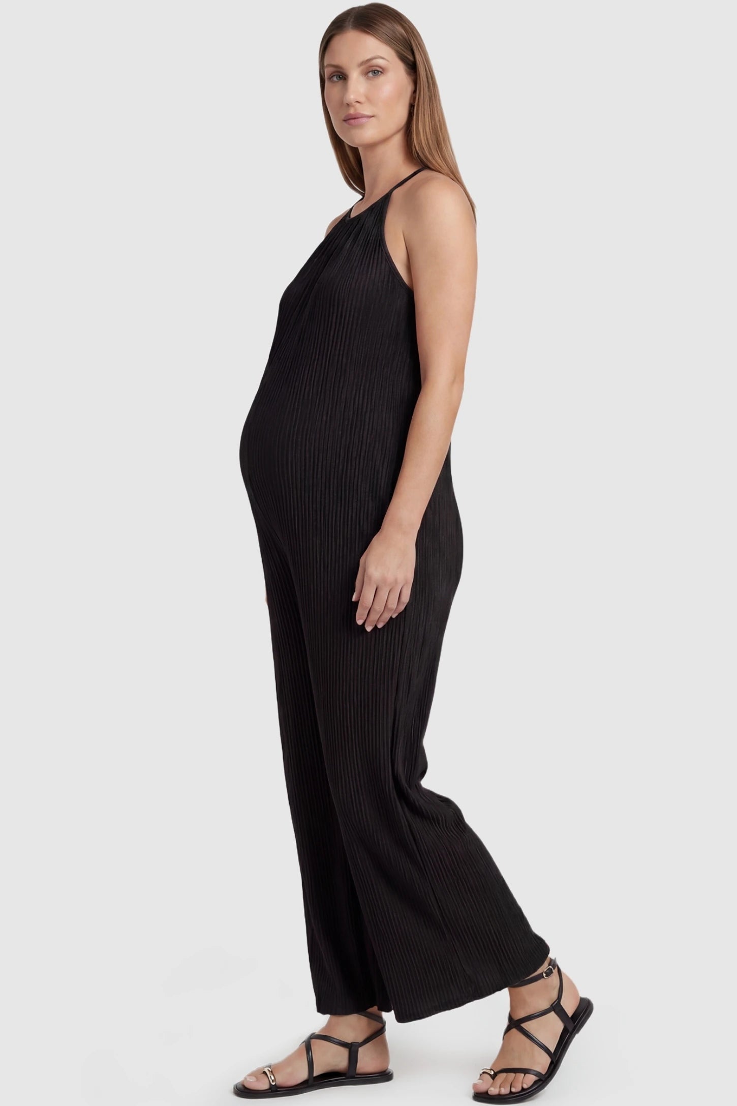 Halter Neck Wide Leg Pocketed Maternity Jumpsuit Halter Neck Wide Leg Pocketed Maternity Jumpsuit in Black featuring a ribbed halter neck design on a pregnant model, side view.