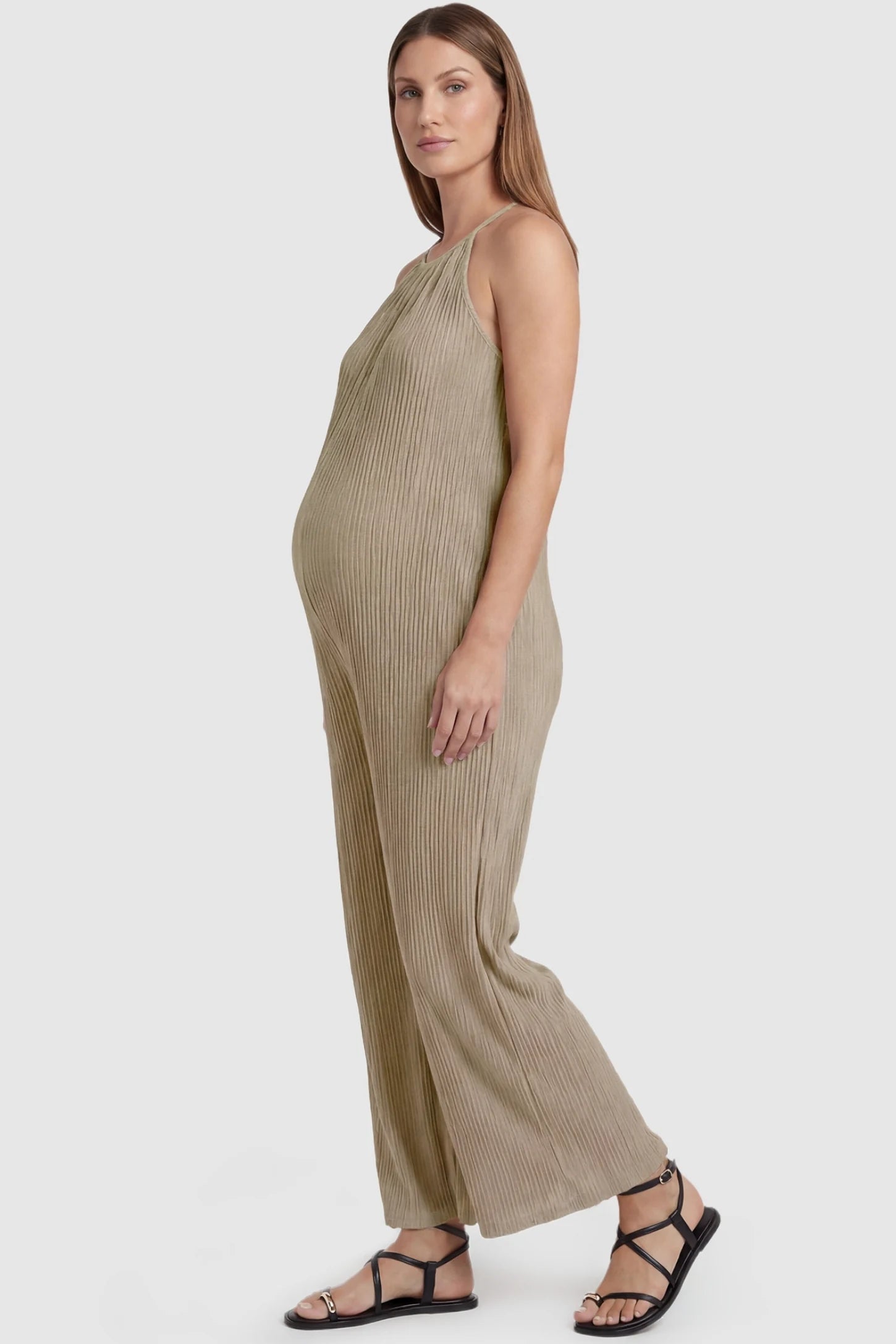 Halter Neck Wide Leg Pocketed Maternity Jumpsuit Halter Neck Wide Leg Pocketed Maternity Jumpsuit in Khaki featuring a ribbed halter neck design on a pregnant model, side view.