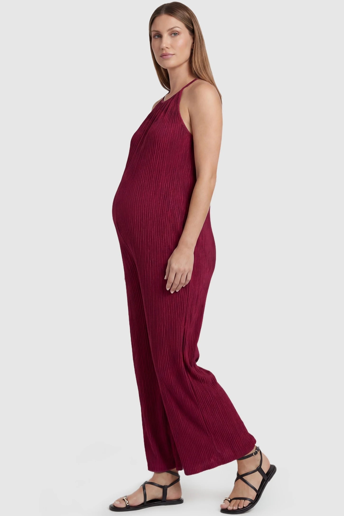 Halter Neck Wide Leg Pocketed Maternity Jumpsuit Halter Neck Wide Leg Pocketed Maternity Jumpsuit in Wine Red featuring a ribbed halter neck design on a pregnant model, side view.