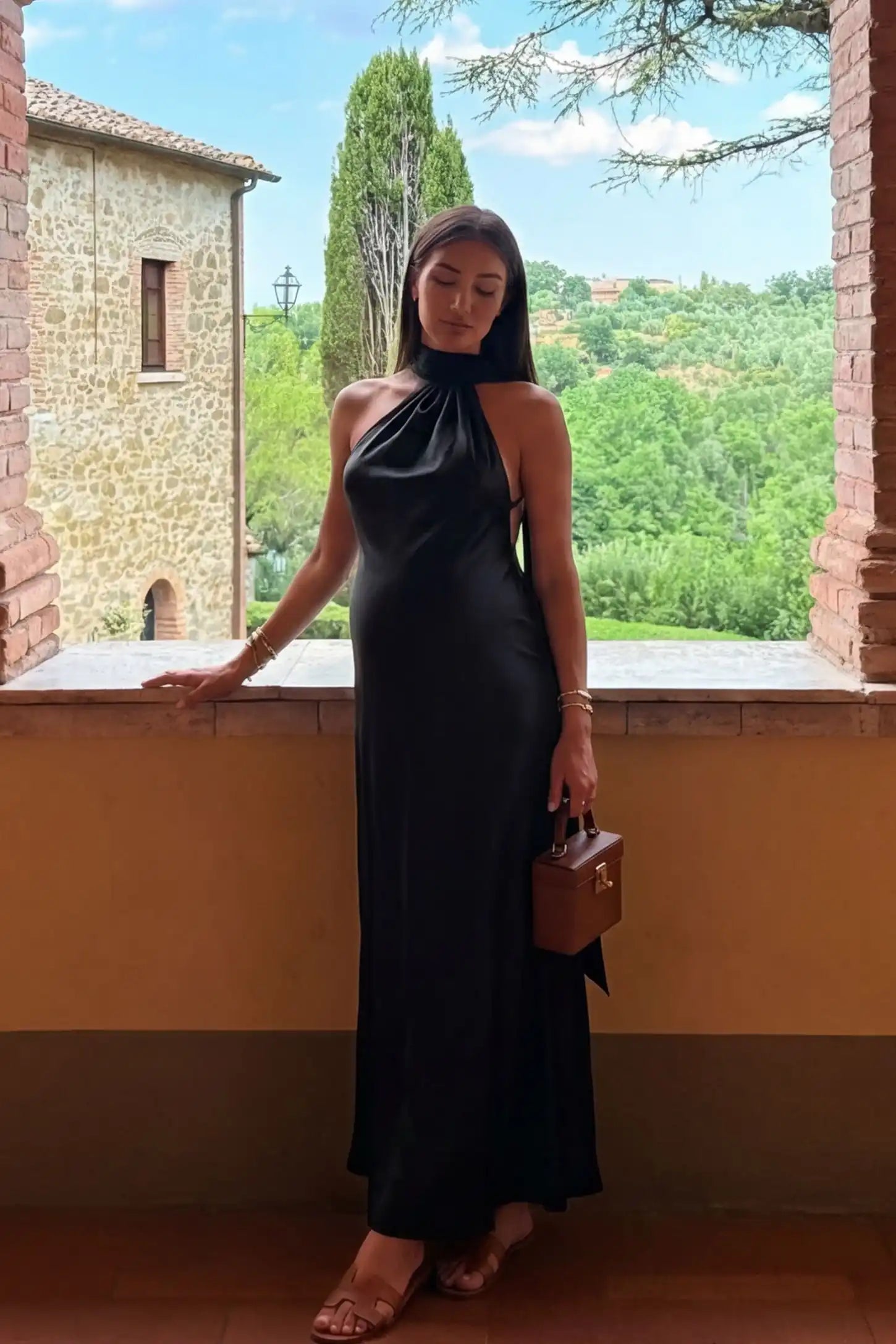 High-Shine Halter Sleeveless Maternity Maxi Dress in Black featuring a floor-length hem on a pregnant model on a balcony.