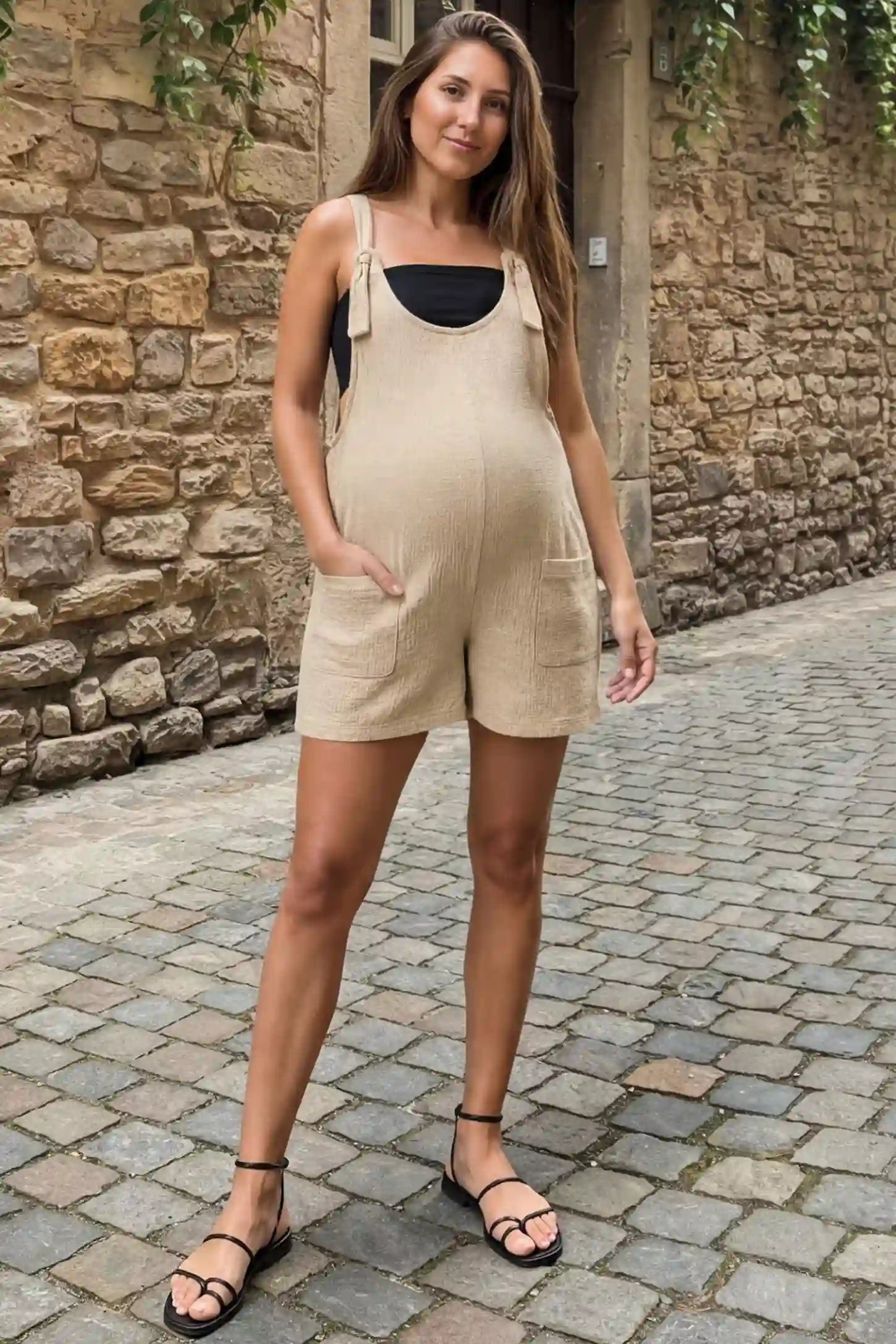 Knot Strap Front Pocket Maternity Romper Knot Strap Front Pocket Maternity Romper in Beige featuring deep front pockets and a scoop neck on a pregnant model walking down a cobblestone street, front view.