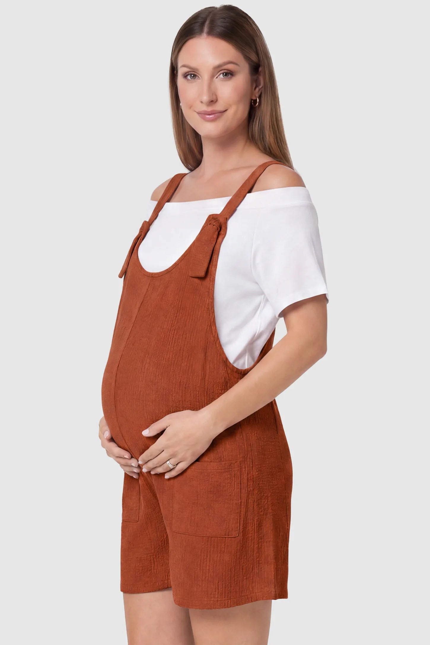Knot Strap Front Pocket Maternity Romper Knot Strap Front Pocket Maternity Romper in Dark Brown featuring deep front pockets and a scoop neck on a pregnant model, side view.