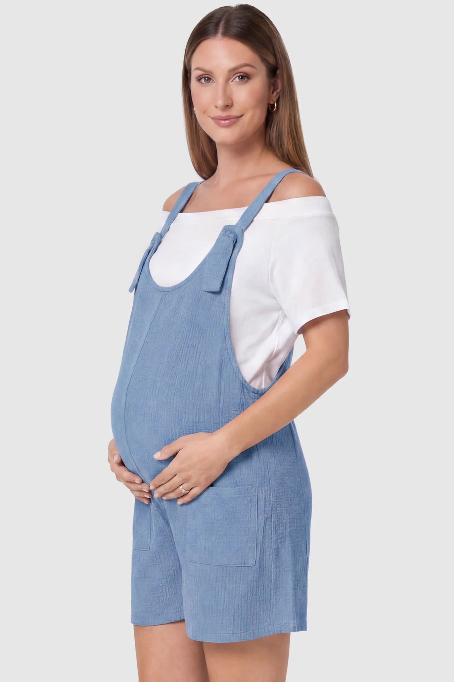 Knot Strap Front Pocket Maternity Romper Knot Strap Front Pocket Maternity Romper in Light Blue featuring deep front pockets and a scoop neck on a pregnant model, side view.
