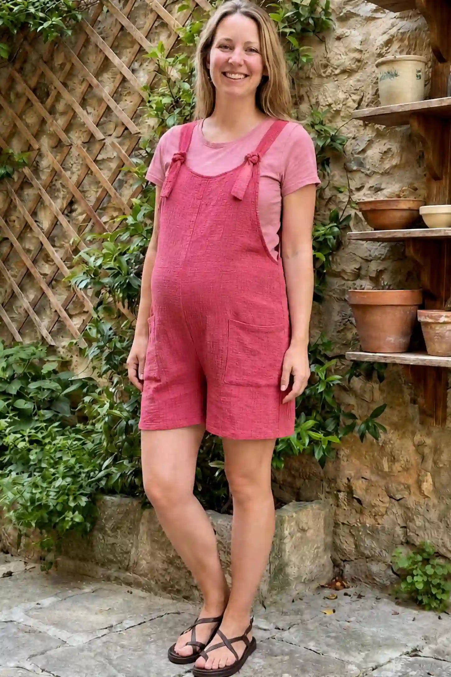 Knot Strap Front Pocket Maternity Romper Knot Strap Front Pocket Maternity Romper in Rose Red featuring a sleeveless scoop-neck design and deep square pockets on a pregnant woman walking down a stone path next to a garden wall, front view.