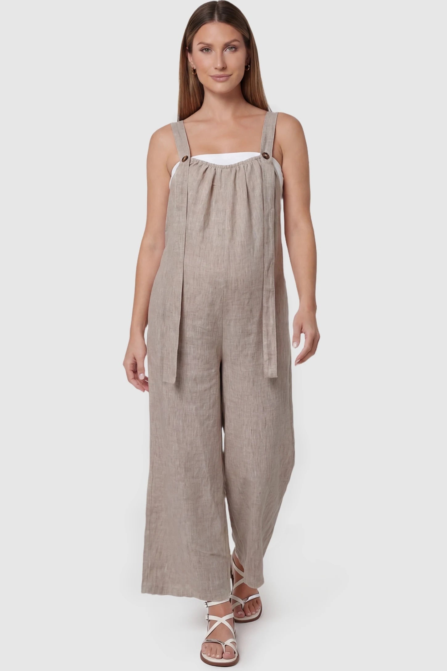Loose Fit Wide Leg Sleeveless Maternity Jumpsuit Loose Fit Wide Leg Sleeveless Maternity Jumpsuit in Light Brown featuring wide shoulder straps with button detail on a pregnant model, front view.