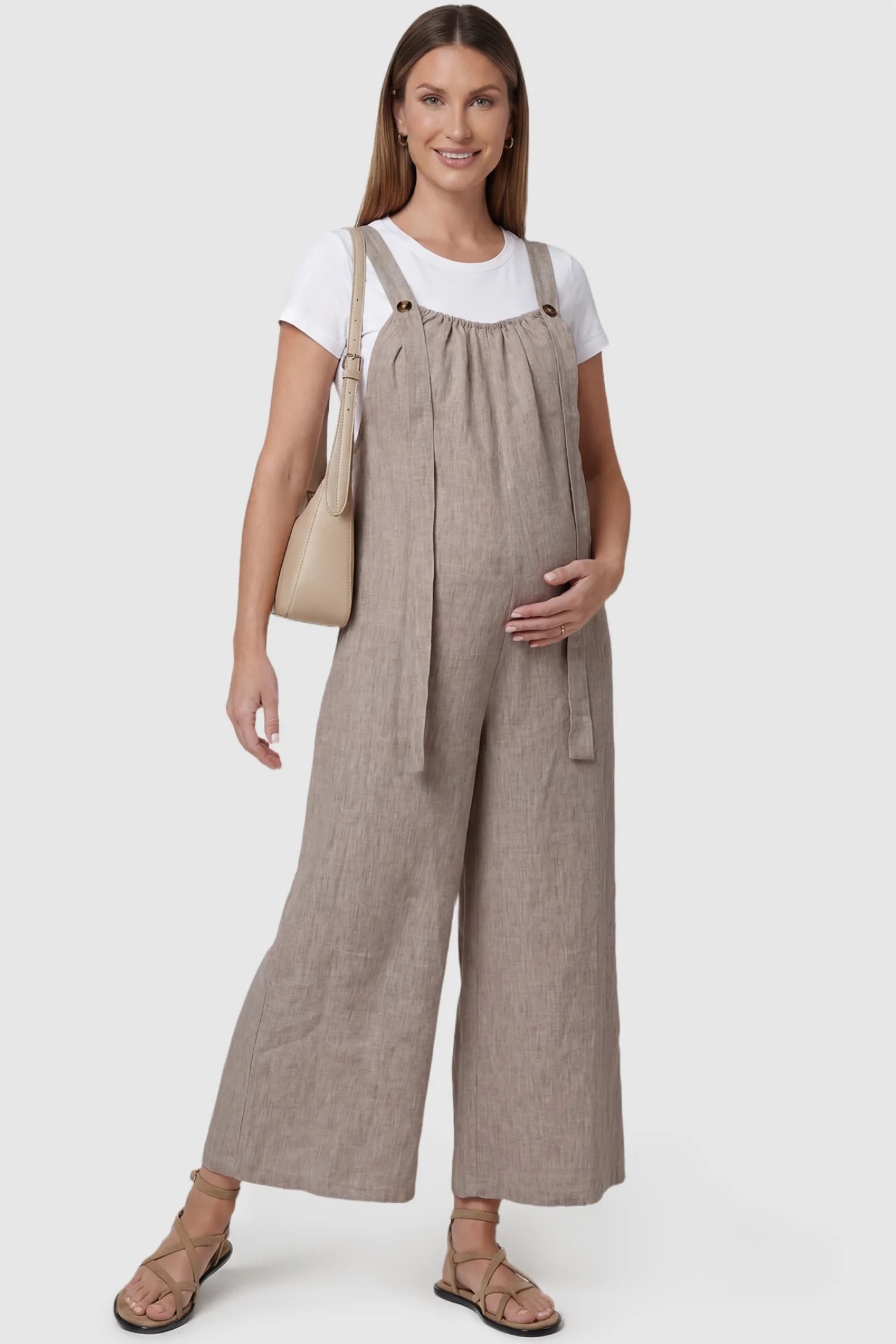 Loose Fit Wide Leg Sleeveless Maternity Jumpsuit Loose Fit Wide Leg Sleeveless Maternity Jumpsuit in Light Brown featuring wide shoulder straps with button detail on a pregnant model, front view.