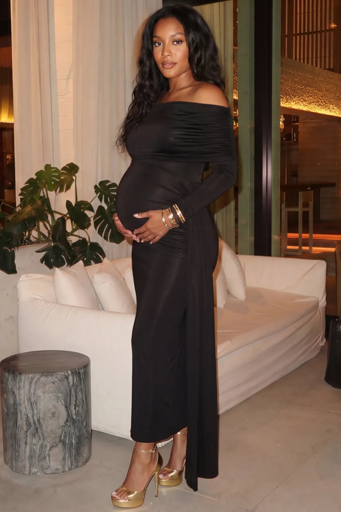 Off Shoulder Draped Wrap Bodycon Maternity Maxi Dress Off Shoulder Draped Wrap Bodycon Maternity Maxi Dress in Black featuring side ruching and long sleeves on a pregnant model cradling her baby bump at an evening party, side view.