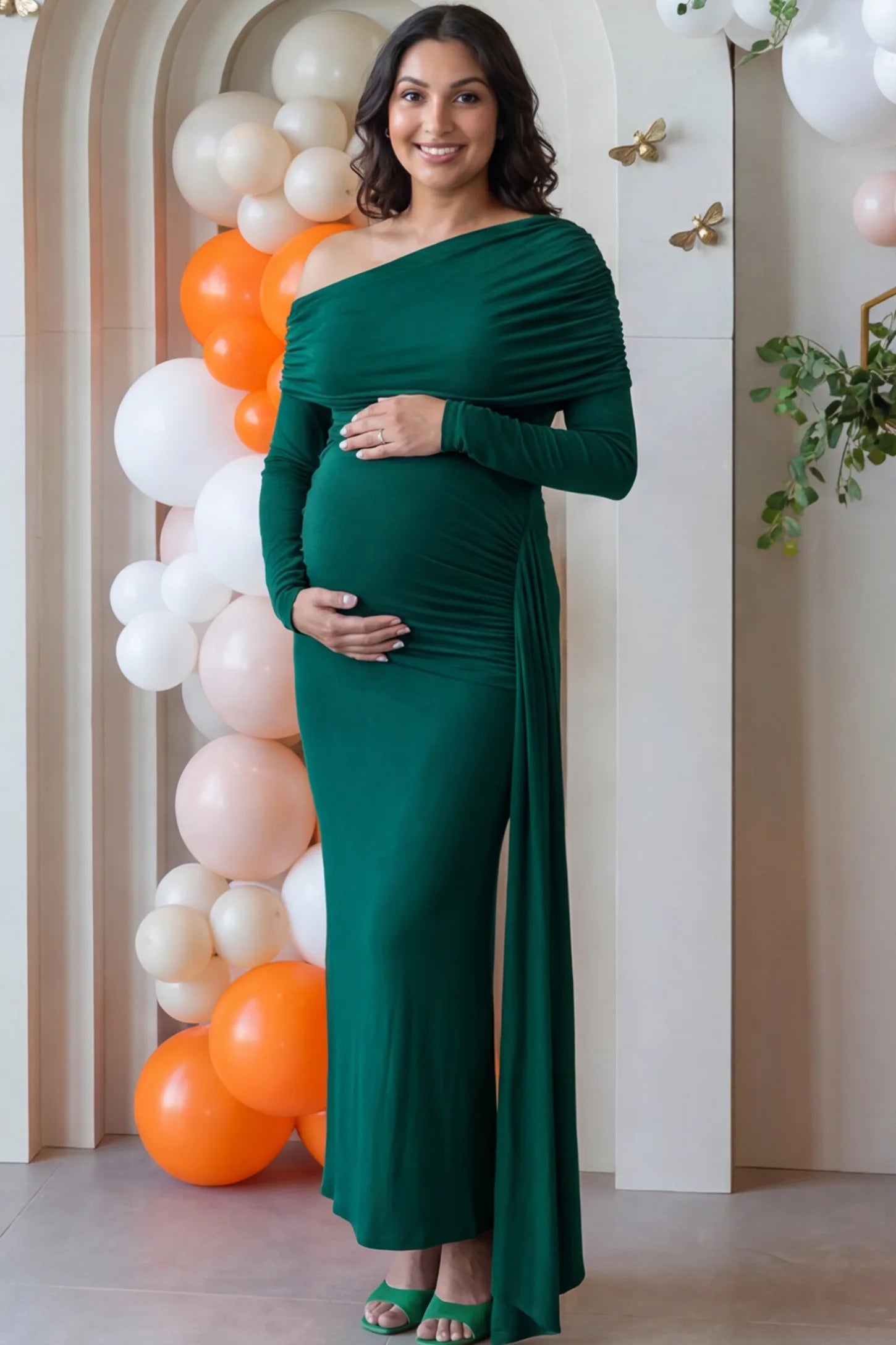 Off Shoulder Draped Wrap Bodycon Maternity Maxi Dress Off Shoulder Draped Wrap Bodycon Maternity Maxi Dress in Green featuring side ruching and long sleeves on a pregnant woman cradling her baby bump at a baby shower, front view.