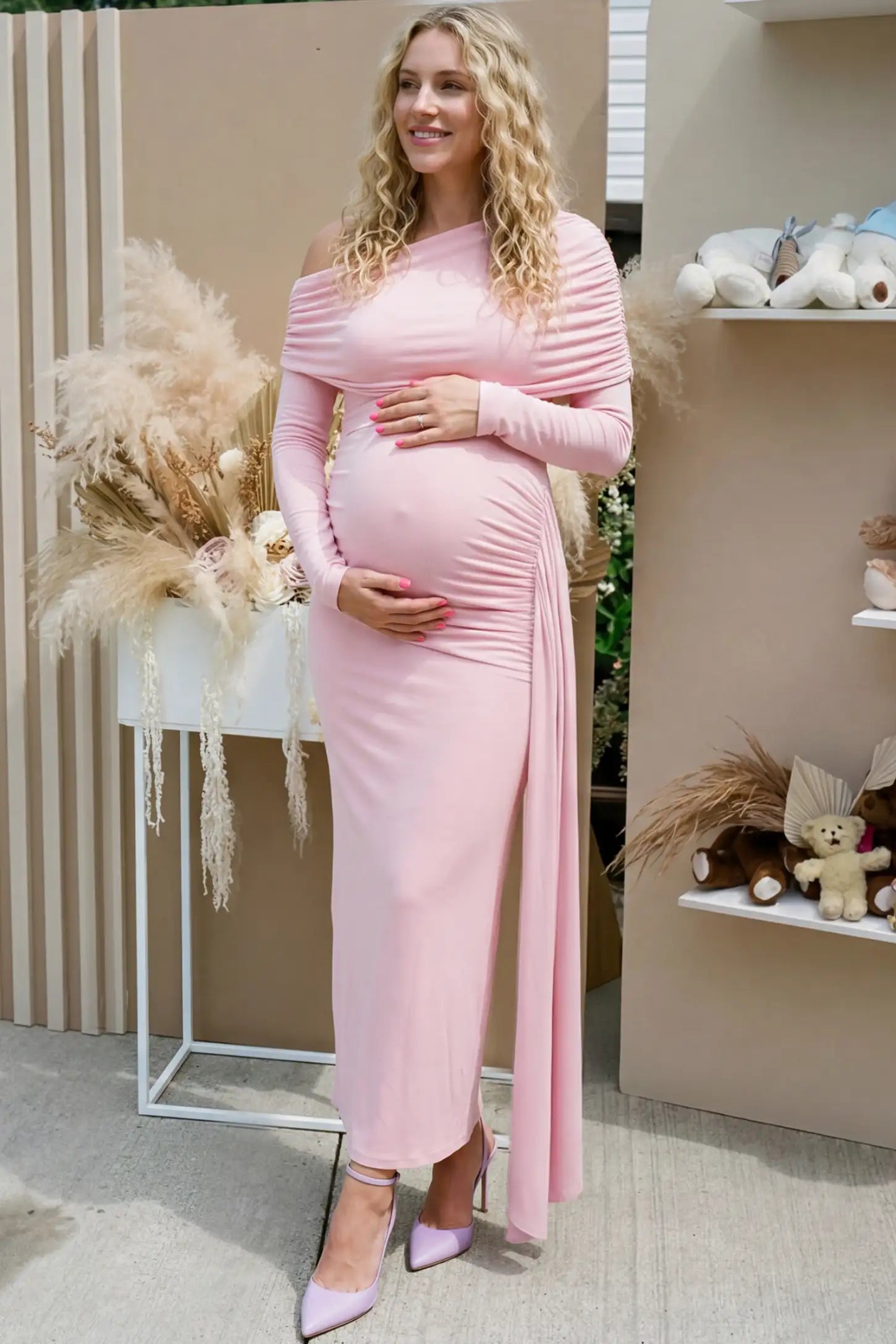 Off Shoulder Draped Wrap Bodycon Maternity Maxi Dress Off Shoulder Draped Wrap Bodycon Maternity Maxi Dress in Pink featuring side ruching and long sleeves on a pregnant model cradling her baby bump at a baby shower, side view.