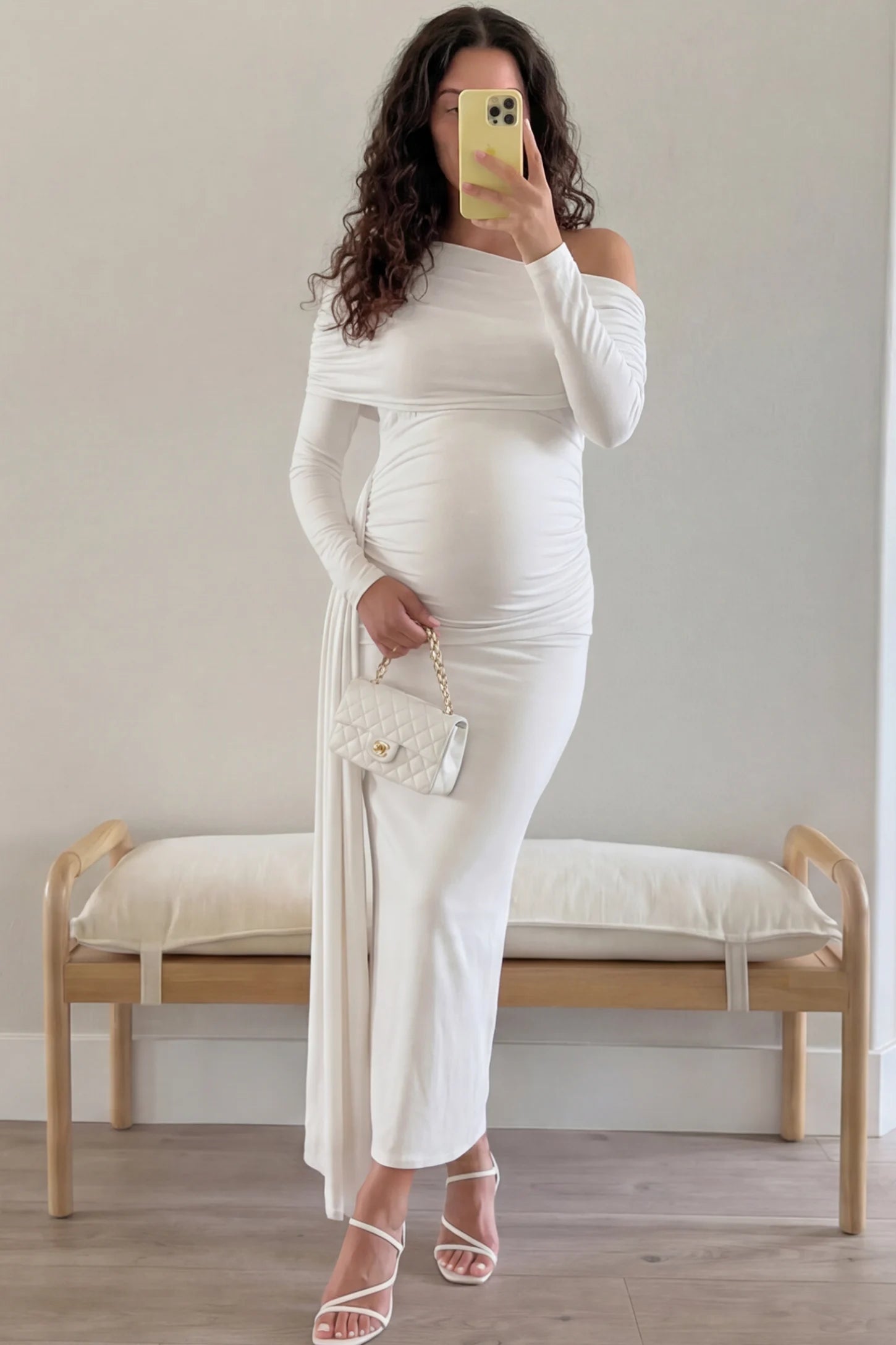 Off Shoulder Draped Wrap Bodycon Maternity Maxi Dress Off Shoulder Draped Wrap Bodycon Maternity Maxi Dress in White featuring side ruching and long sleeves on a pregnant woman taking a mirror selfie in a living room, front view.