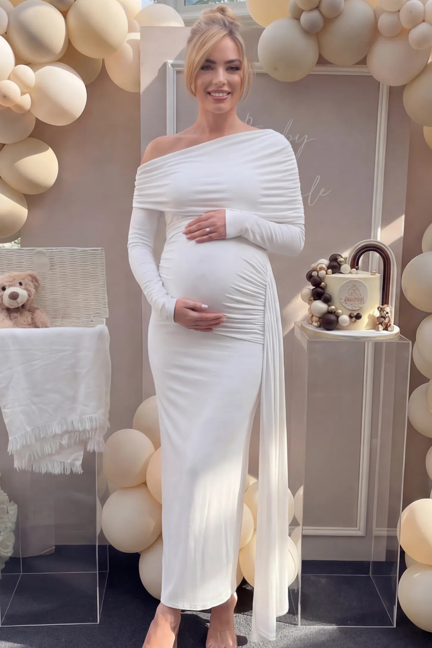 Off Shoulder Draped Wrap Bodycon Maternity Maxi Dress Off Shoulder Draped Wrap Bodycon Maternity Maxi Dress in White featuring side ruching and long sleeves on a pregnant woman cradling her baby bump at a baby shower, front view.