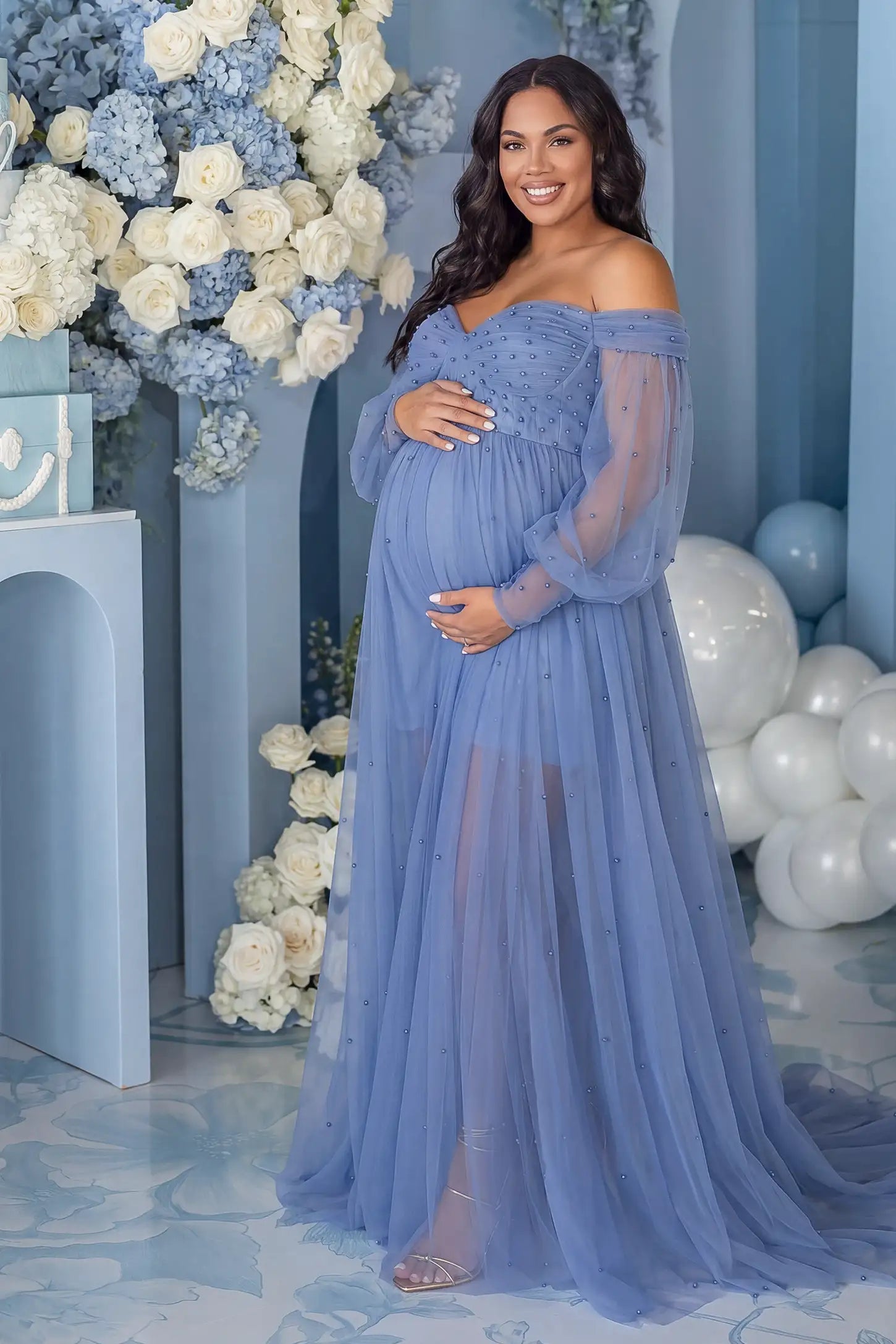 Off-Shoulder Empire Waist Tulle Maternity Gown Off-Shoulder Empire Waist Maternity Gown in Blue featuring an off-shoulder sweetheart neckline, outdoor lifestyle front view on a pregnant model.