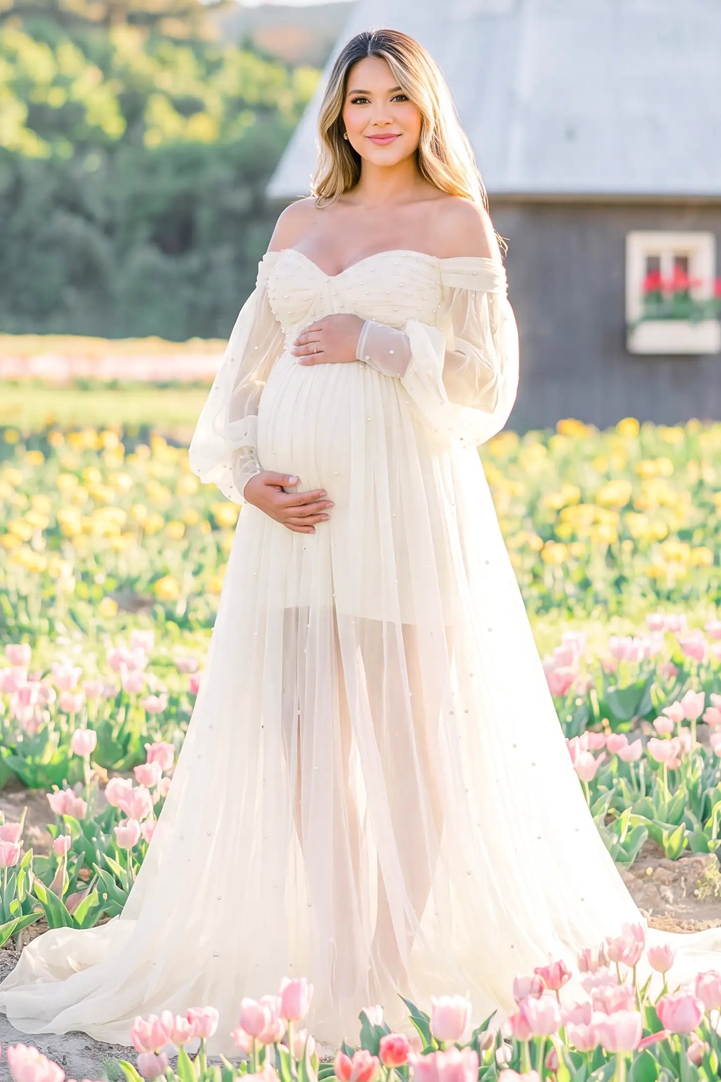 Off-Shoulder Empire Waist Tulle Maternity Gown Off-Shoulder Empire Waist Maternity Gown in Ivory featuring an off-shoulder sweetheart neckline, outdoor lifestyle front view on a pregnant model.