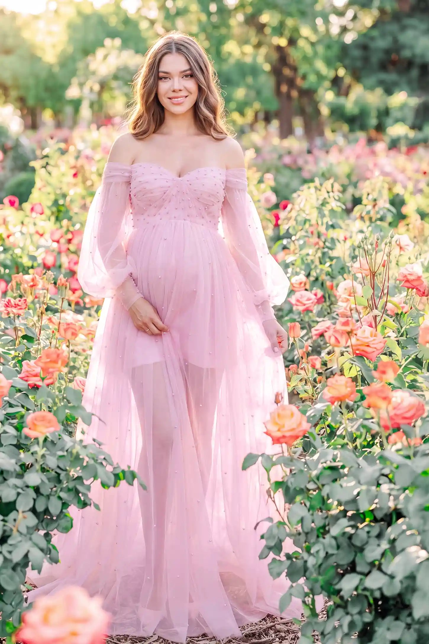 Off-Shoulder Empire Waist Tulle Maternity Gown Off-Shoulder Empire Waist Maternity Gown in Pink featuring an off-shoulder sweetheart neckline, outdoor lifestyle front view on a pregnant model.