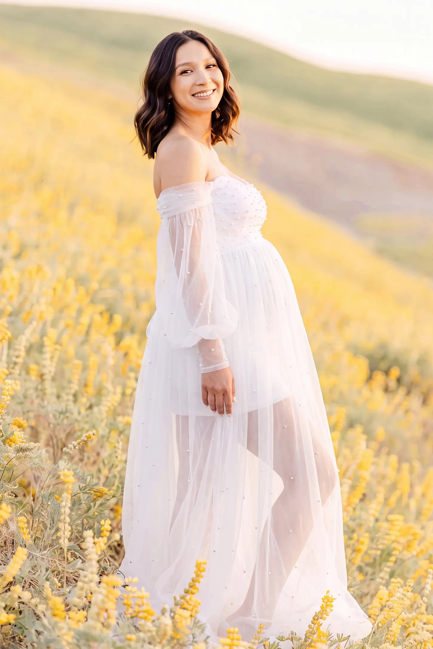 Off-Shoulder Empire Waist Tulle Maternity Gown Off-Shoulder Empire Waist Maternity Gown in White with a flowing A-line shape, outdoor lifestyle view on a pregnant model.
