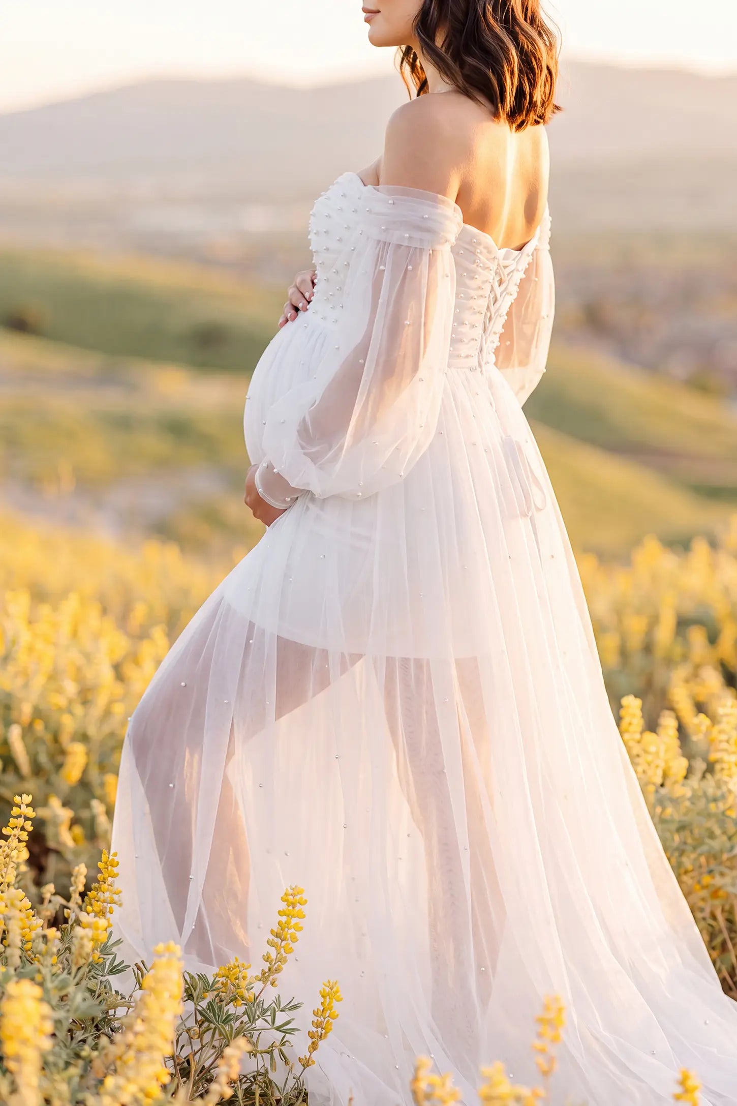 Off-Shoulder Empire Waist Tulle Maternity Gown Off-Shoulder Empire Waist Maternity Gown in White with a flowing A-line shape, outdoor lifestyle view on a pregnant model.