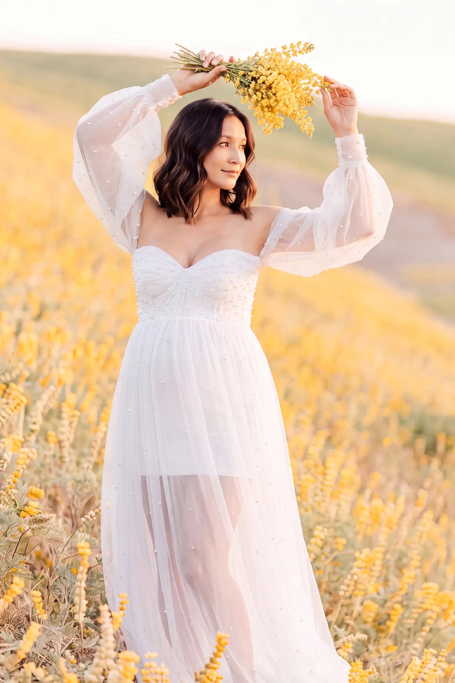 Off-Shoulder Empire Waist Tulle Maternity Gown Off-Shoulder Empire Waist Maternity Gown in White featuring an off-shoulder sweetheart neckline, outdoor lifestyle front view on a pregnant model.