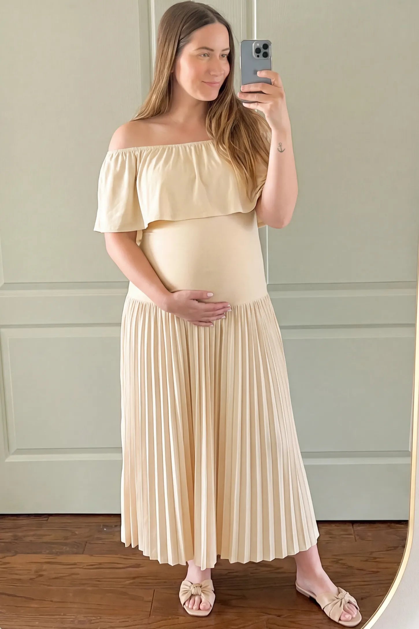 Pleated Off Shoulder Maternity Maxi Dress Pleated Off Shoulder Maternity Maxi Dress in Beige featuring a high stretch pleated maxi skirt on a pregnant model during an indoor setting, front view.