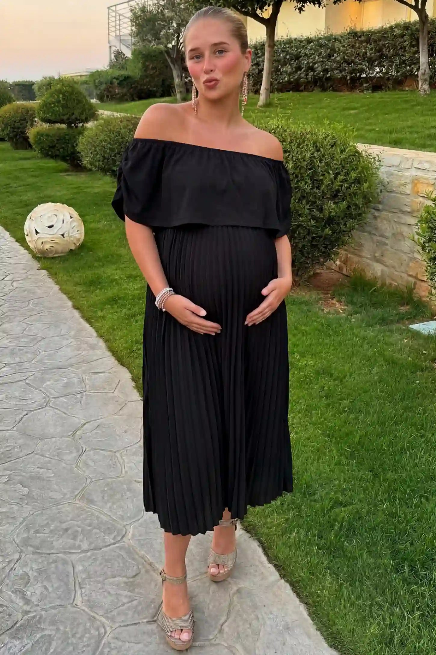 Pleated Off Shoulder Maternity Maxi Dress Pleated Off Shoulder Maternity Maxi Dress in Black featuring a high stretch pleated maxi skirt on a pregnant model during an outdoor photoshoot, front view.