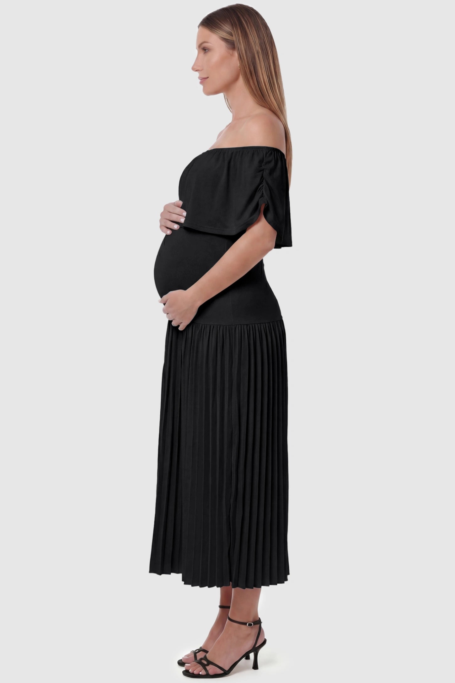 Pleated Off Shoulder Maternity Maxi Dress Pleated Off Shoulder Maternity Maxi Dress  in Black featuring a high stretch pleated maxi skirt on a pregnant model, side view.
