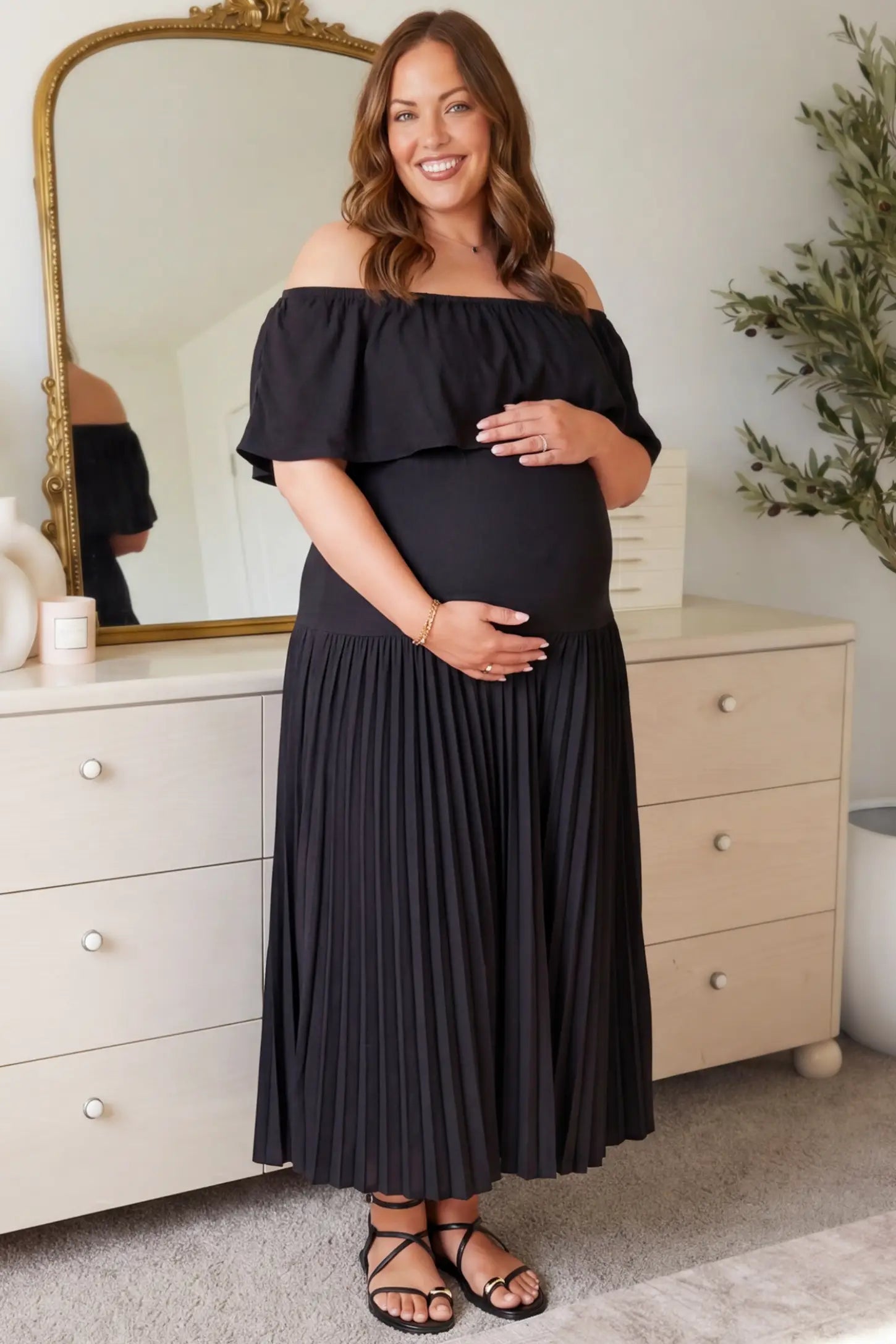 Pleated Off Shoulder Maternity Maxi Dress Pleated Off Shoulder Maternity Maxi Dress in Black featuring a high stretch pleated maxi skirt on a pregnant model during an indoor setting, front view.