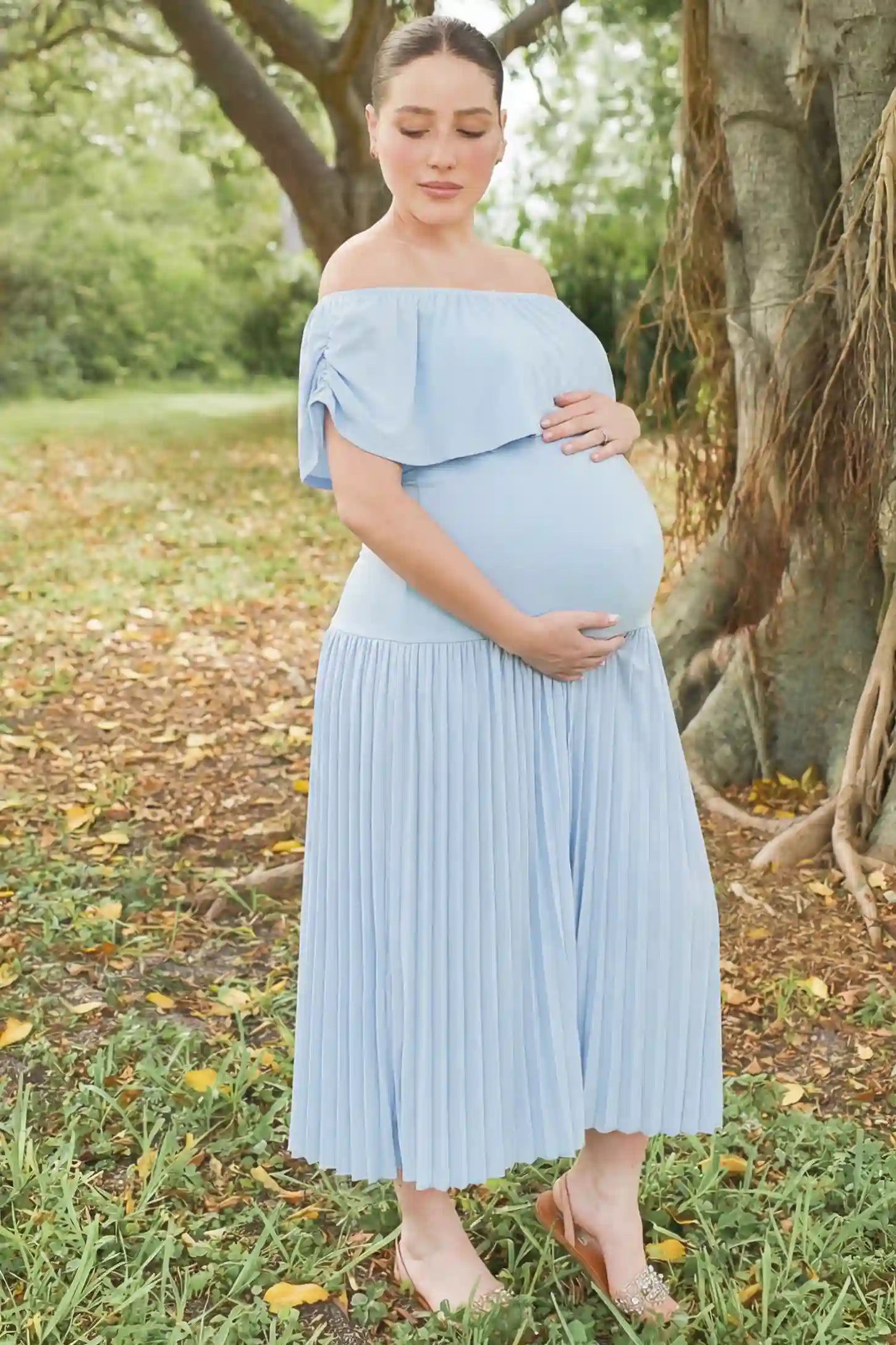 Pleated Off Shoulder Maternity Maxi Dress Pleated Off Shoulder Maternity Maxi Dress in Light Blue featuring a high stretch pleated maxi skirt on a pregnant model during an outdoor photoshoot, side view.