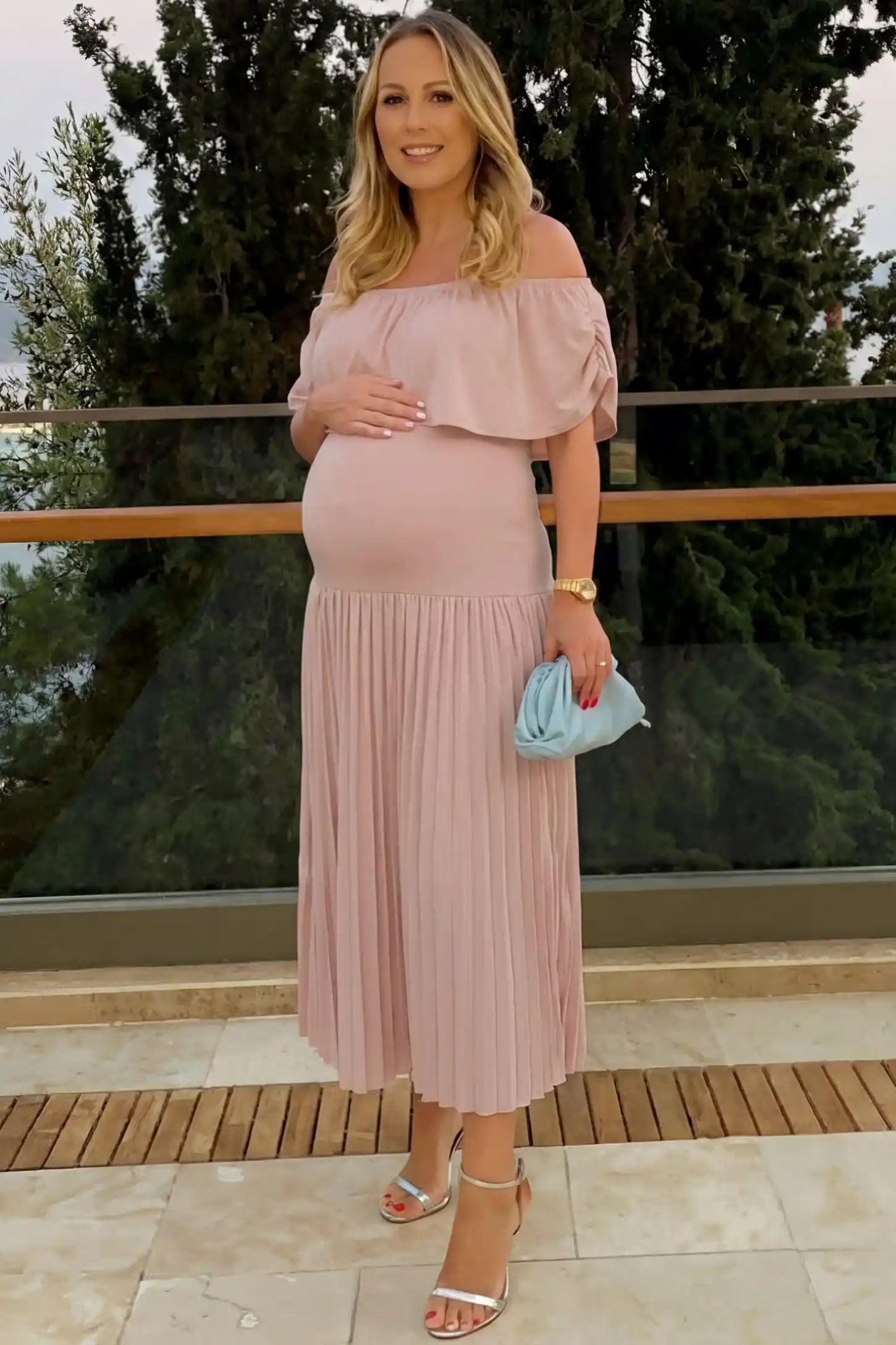 Pleated Off Shoulder Maternity Maxi Dress Pleated Off Shoulder Maternity Maxi Dress in Pink featuring a high stretch pleated maxi skirt on a pregnant model during an outdoor photoshoot, front view.